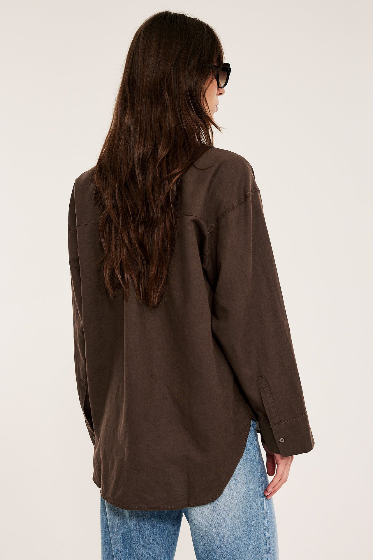 Widok z tyłu: kobieta w luźnej, ciemnobrązowej lnianej koszuli, jasnoniebieskich jeansach i okularach.