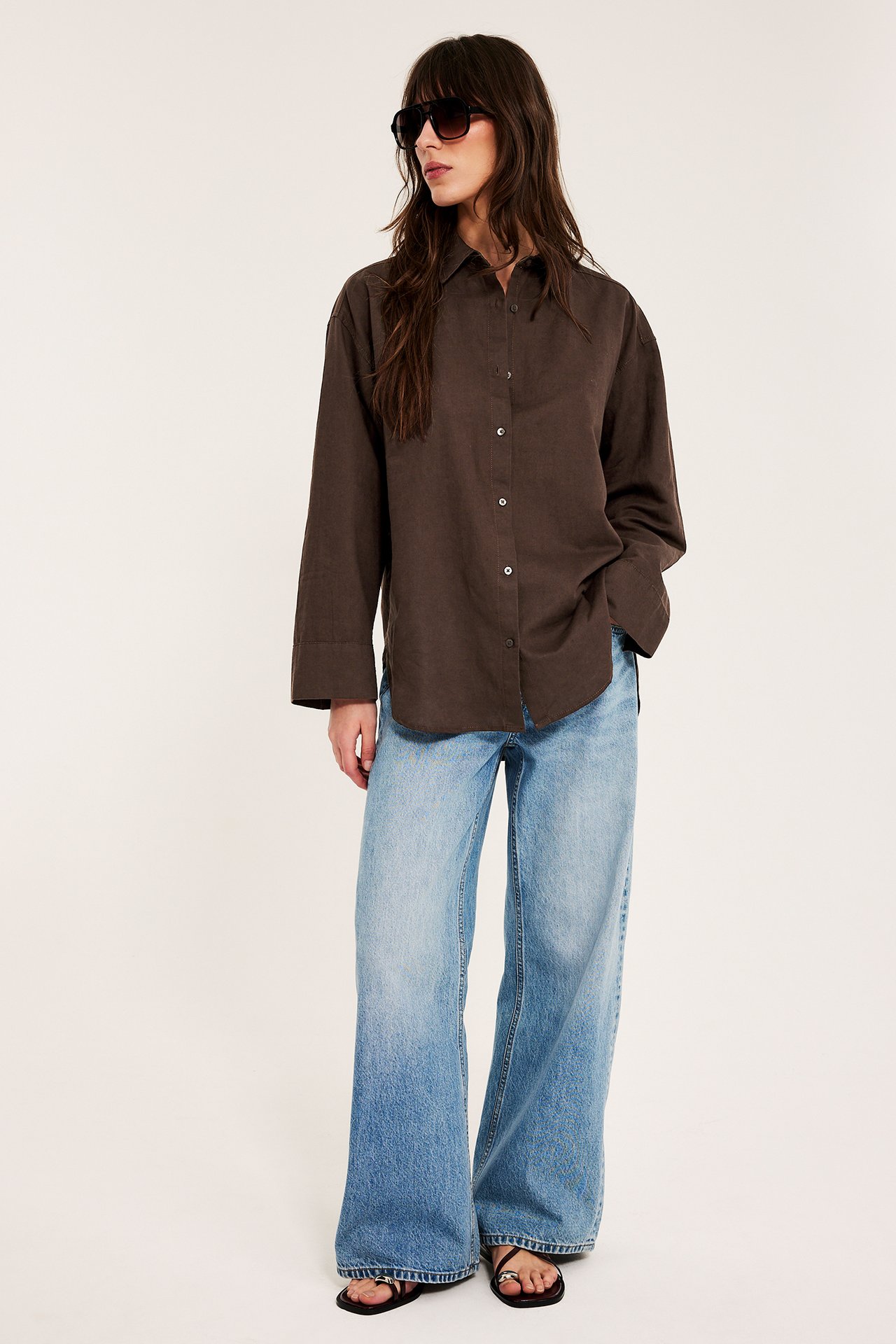 Kvinne i oversized mørkebrun lin skjorte, lyseblå vide jeans, svarte sandaler og solbriller.