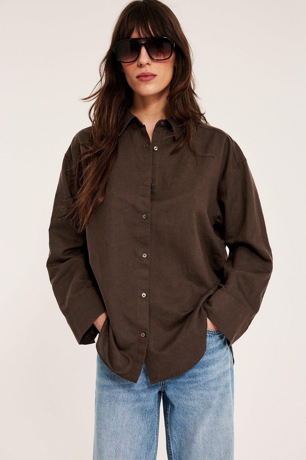 Z przodu: kobieta w ciemnobrązowej lnianej koszuli, jasnoniebieskich jeansach i czarnych okularach.