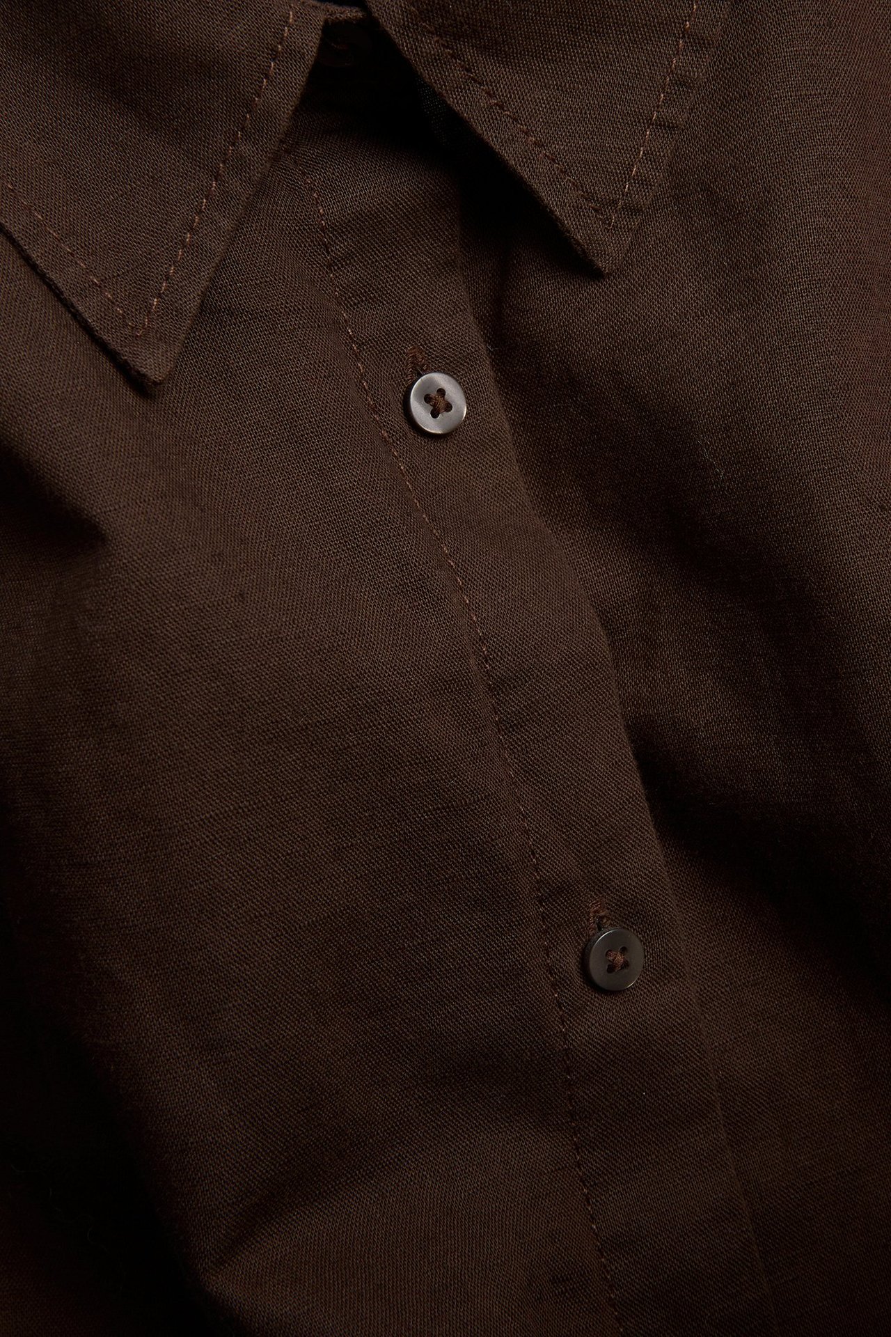 Lähikuva tummanruskeasta ylisuuresta pellavasekoitepaidasta, jossa näkyy kaulus ja nappilista yhteensopivine nappeineen.