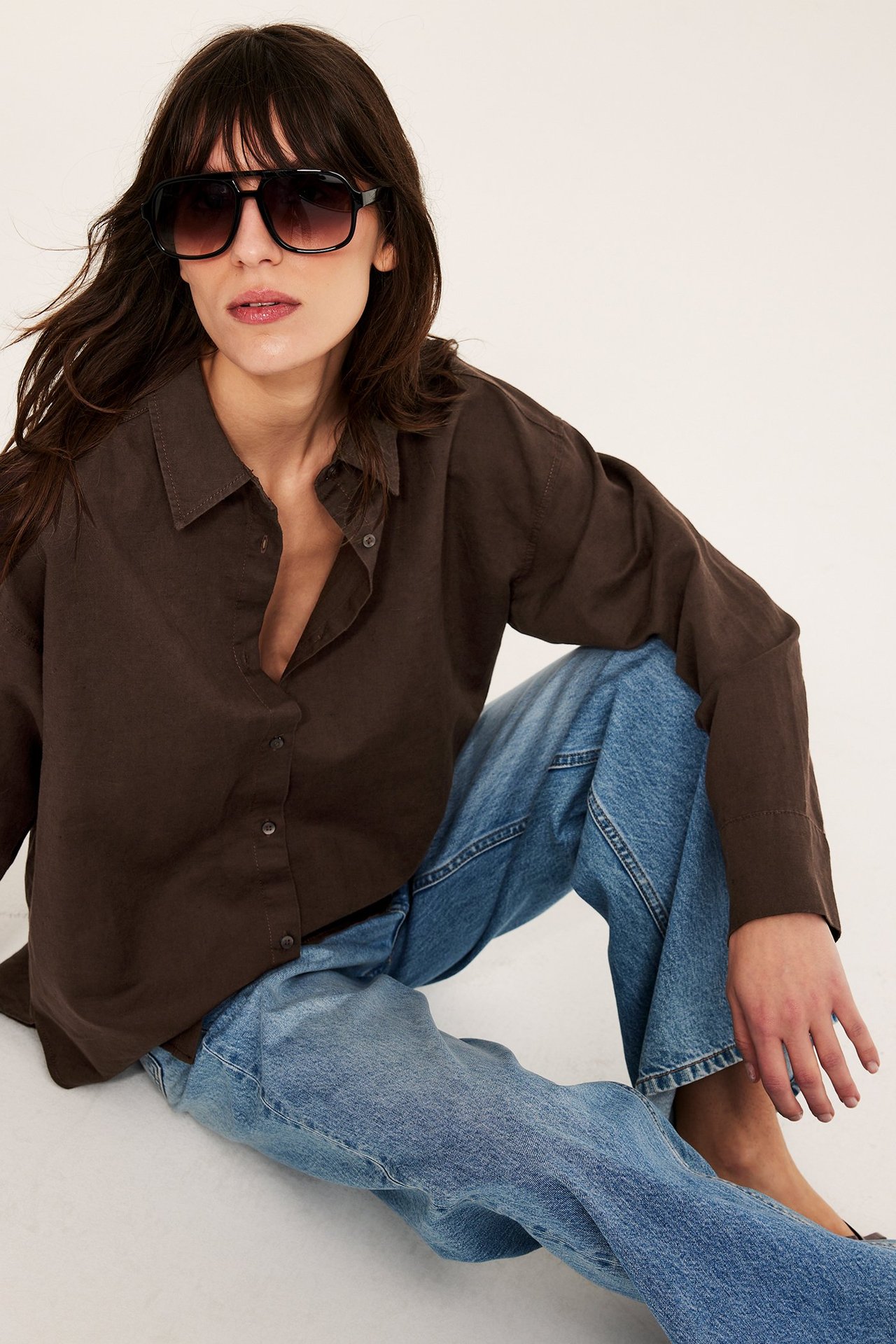 Forfra: Kvinne i oversized mørk brun linblanding skjorte, lyseblå jeans og svarte solbriller.