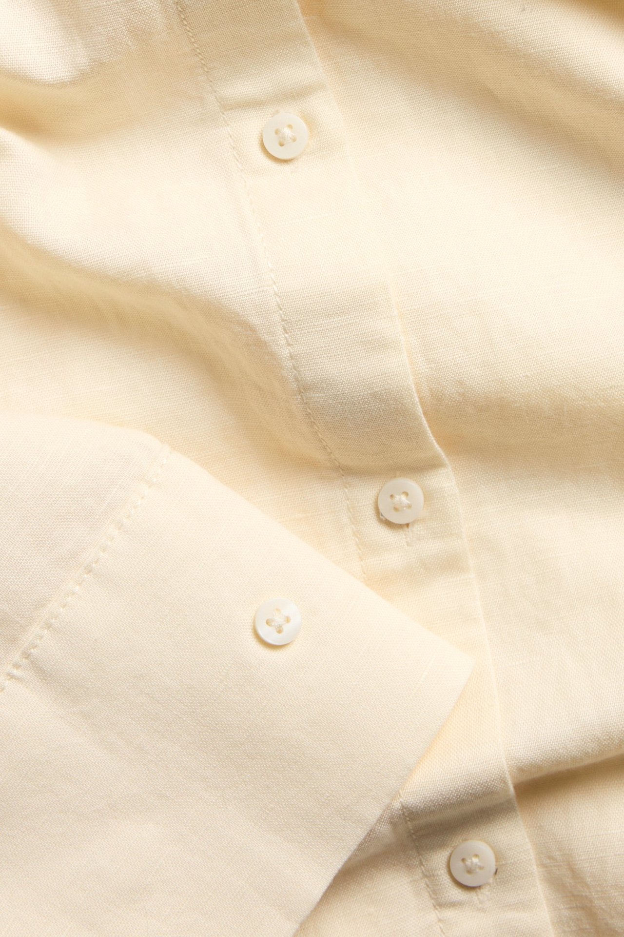 Zbliżenie na jasnożółtą koszulę z mieszanki lnu, ukazujące guziki na plisie i mankiecie.