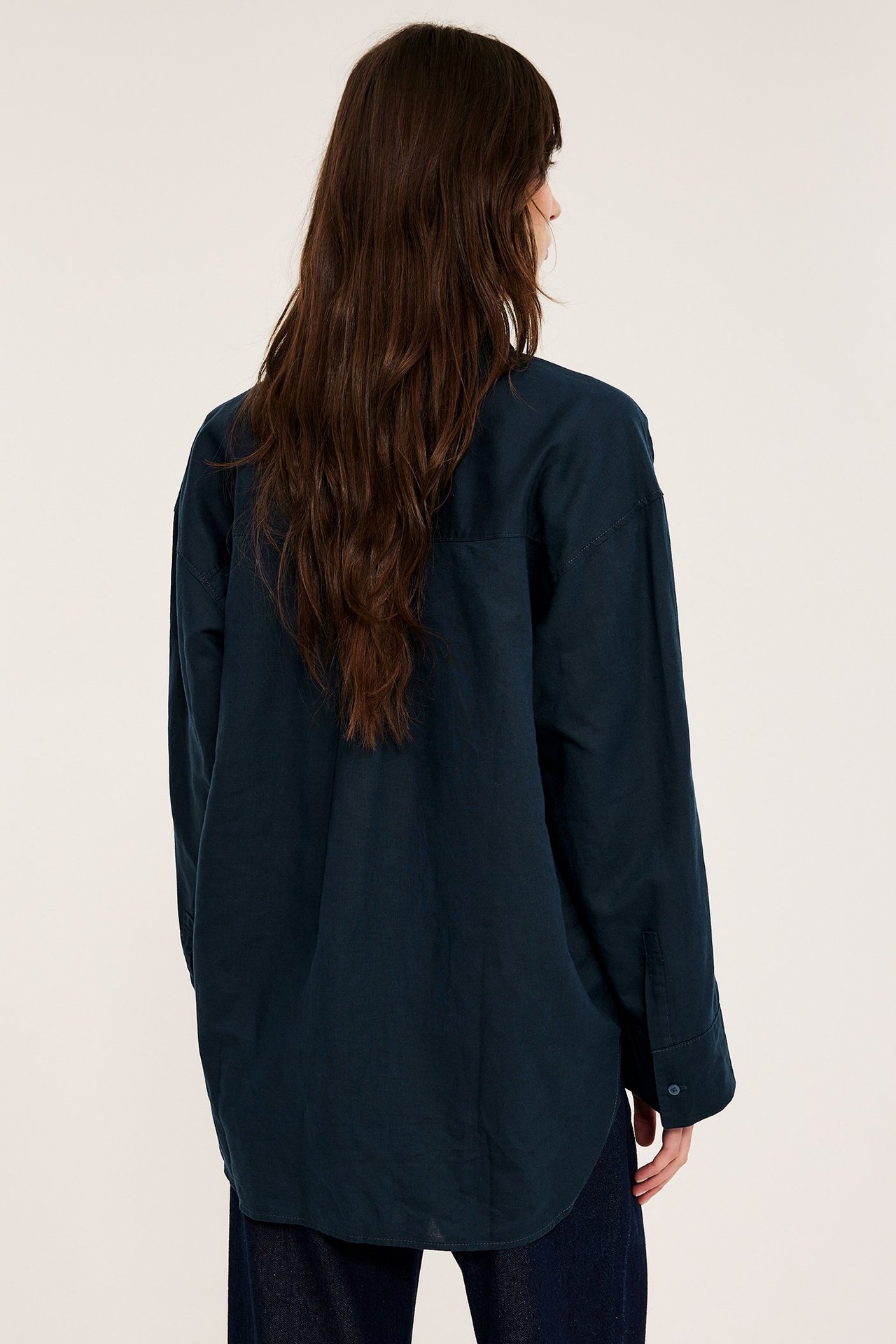 Tył kobiety w ciemnoniebieskiej, oversize koszuli lnianej z długimi rękawami, na ciemnych spodniach.