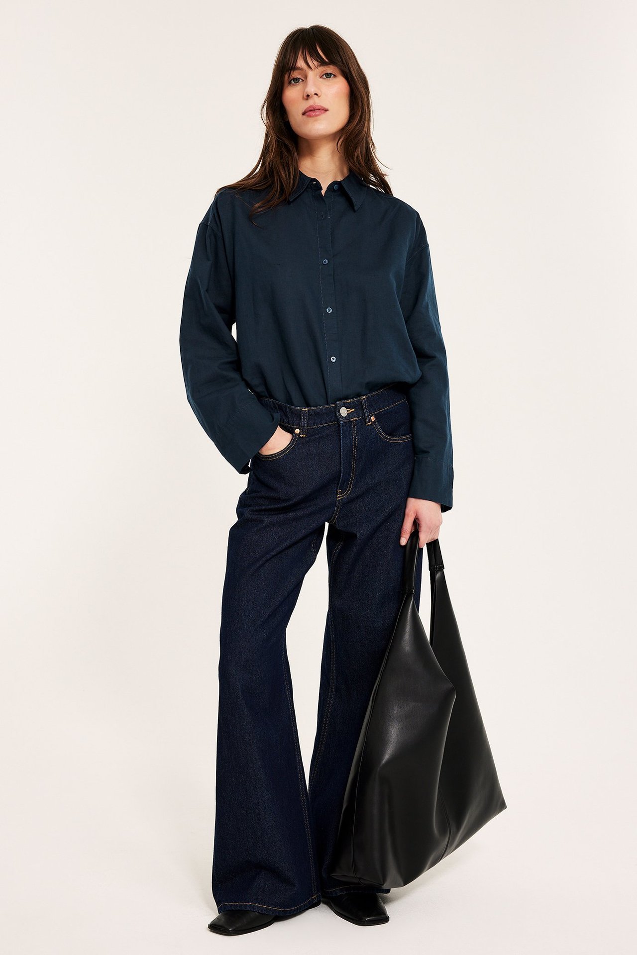 Kvinne i mørk blå oversized skjorte i linblanding, mørke vide jeans, holder svart totebag, forfra.