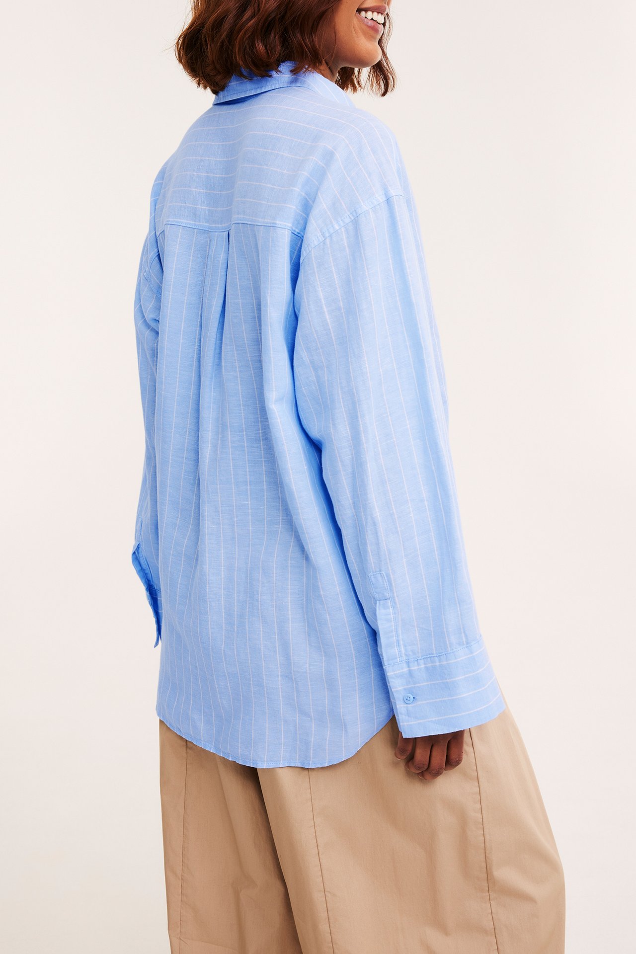Bakifrån: kvinna i en oversized ljusblå randig skjorta i linneblandning och ljusbeige byxor.