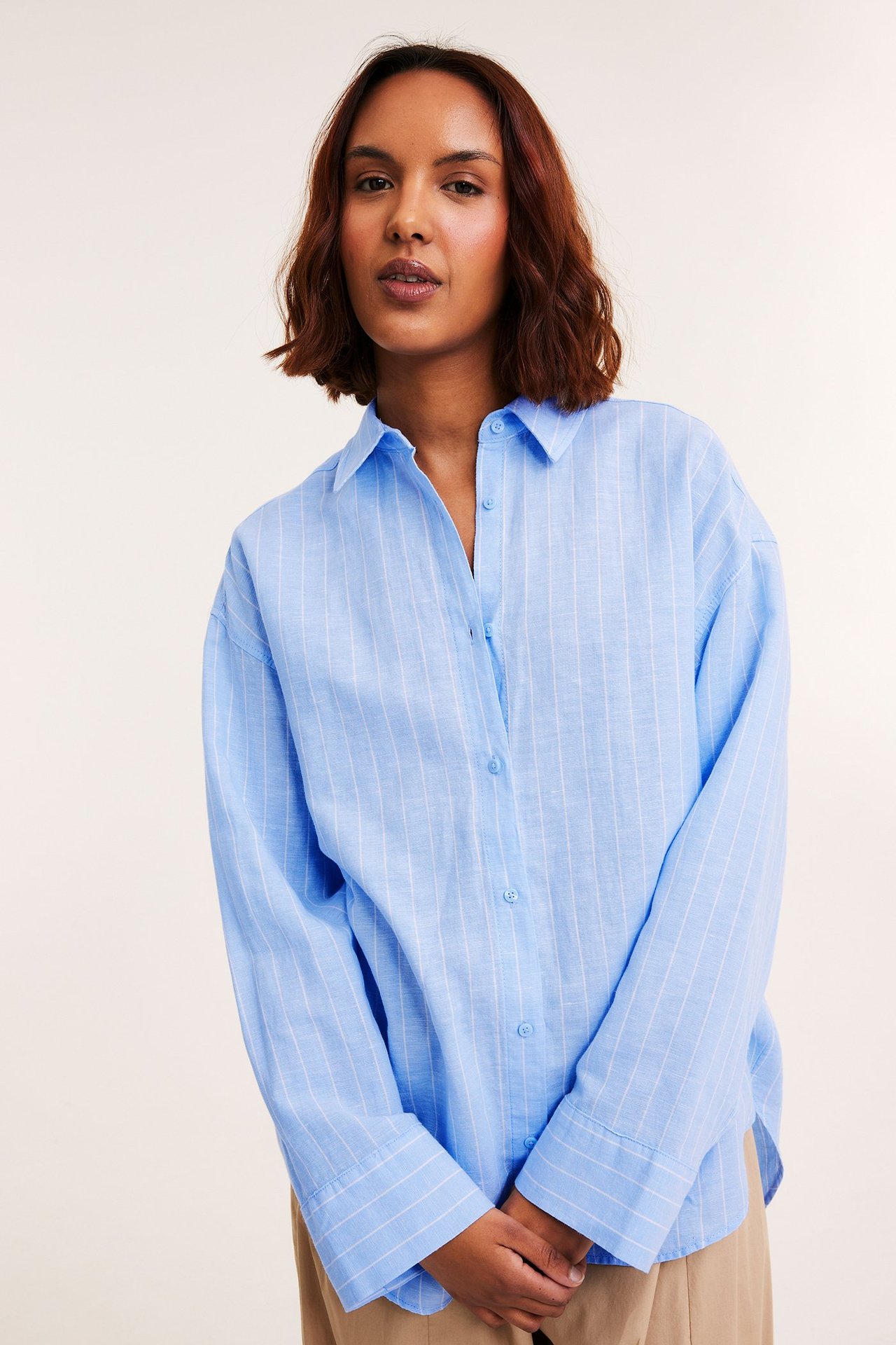 Framifrån, kvinna i oversize ljusblå randig linneskjorta och beiga byxor.