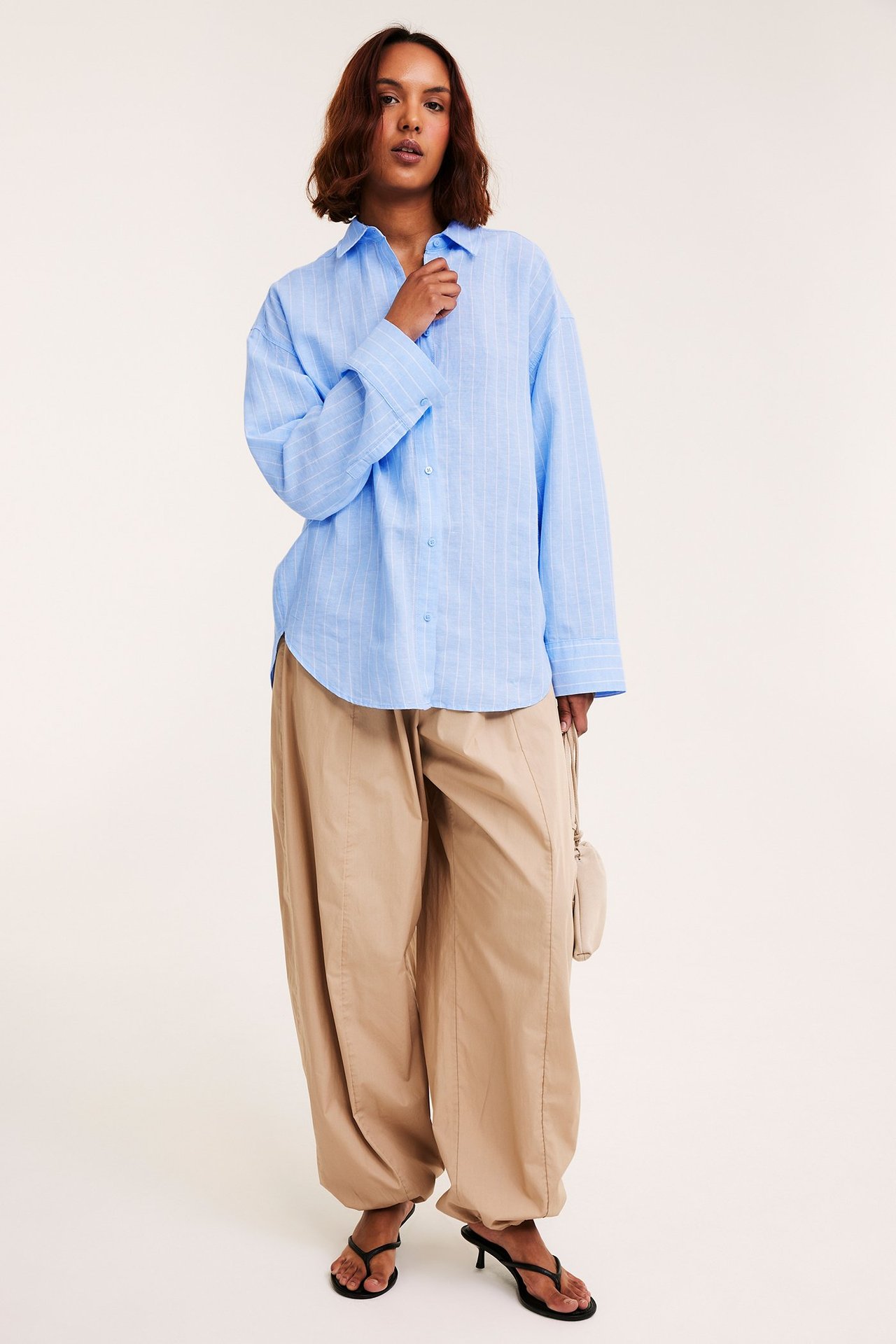 Widok z przodu: kobieta w luźnej błękitnej koszuli w paski, beżowych szerokich spodniach, czarnych sandałach, trzyma beżową torebkę.