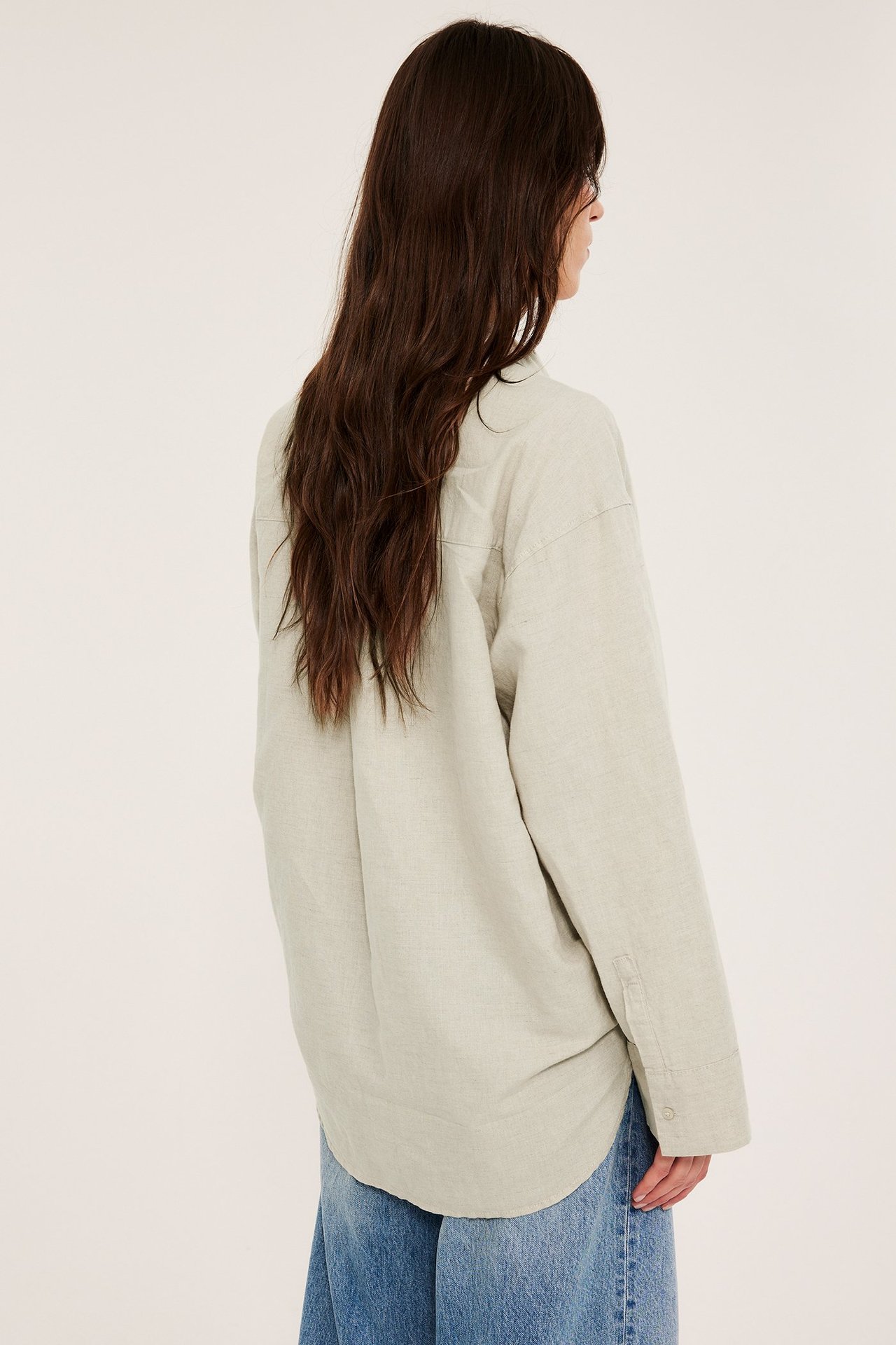 Widok z tyłu kobiety w obszernej, beżowej melanżowej lnianej koszuli i niebieskich dżinsach z szerokimi nogawkami.