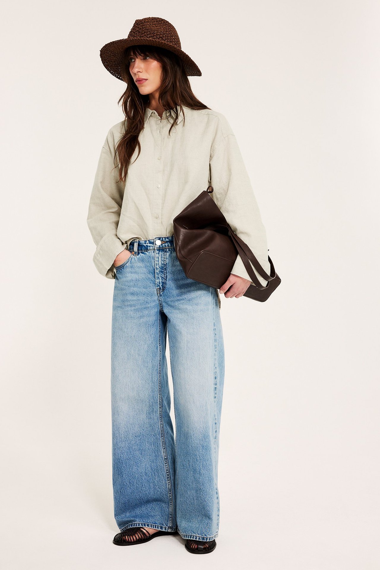 Forfra: kvinne i oversized beigemelert linskjorte, vide jeans, mørkebrun stråhatt, og holder en brun veske.