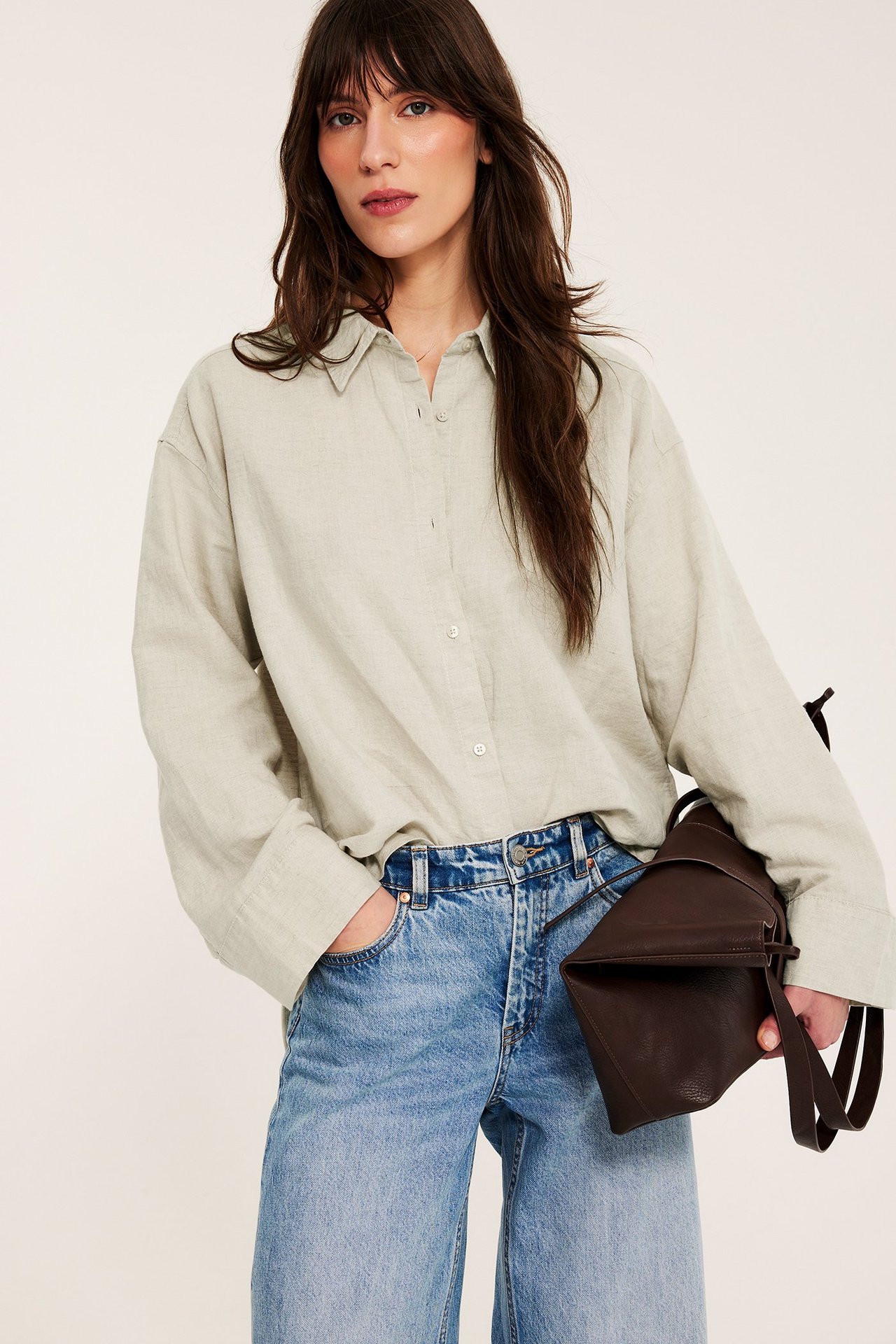 Kobieta przodem w oversize'owej jasnobeżowej koszuli z lnu, niebieskich szerokich dżinsach, trzyma brązową torebkę.