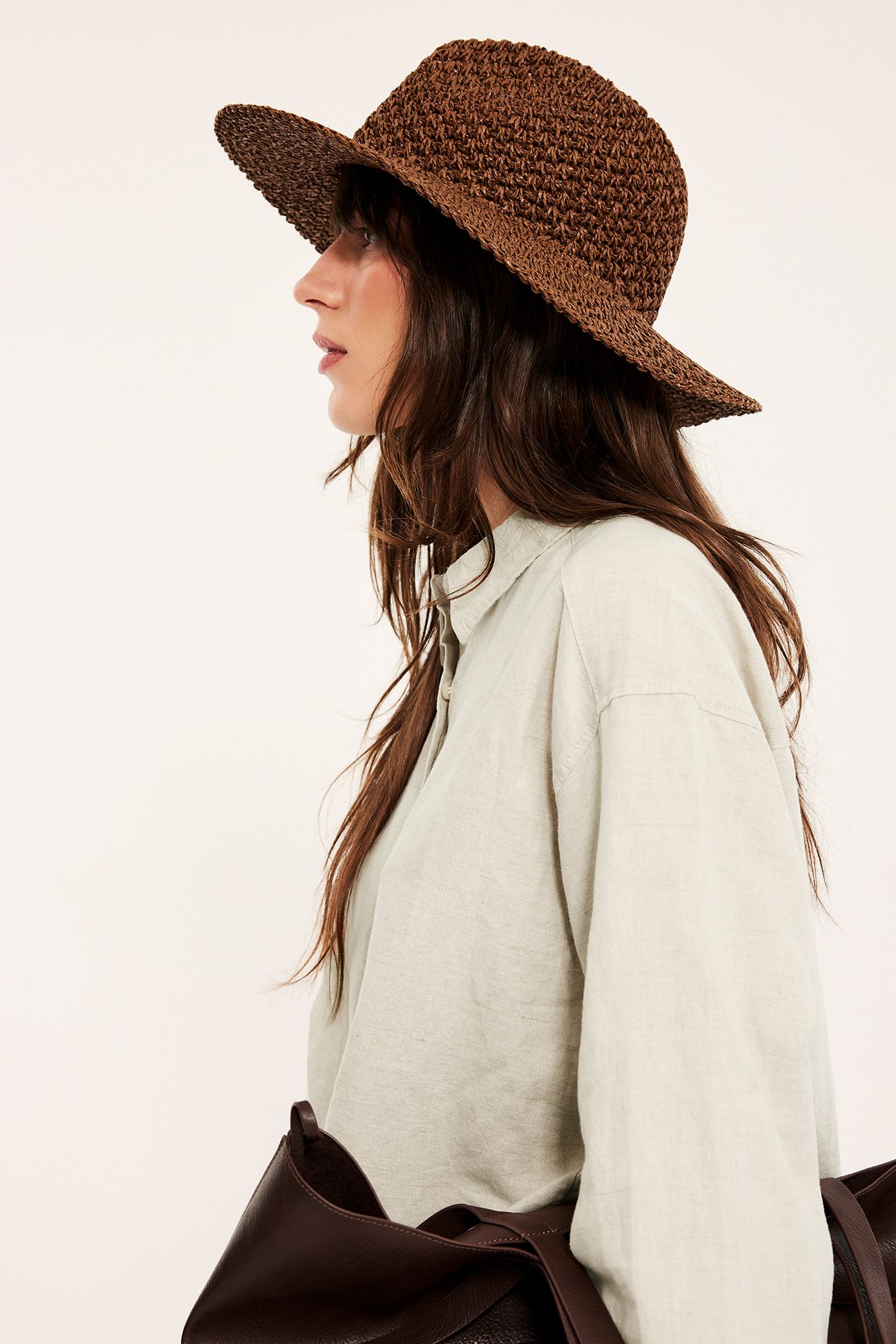 Sidevisning: Kvinne i oversized beigemelert linskjorte, brun vevd hatt, bærer brun veske.