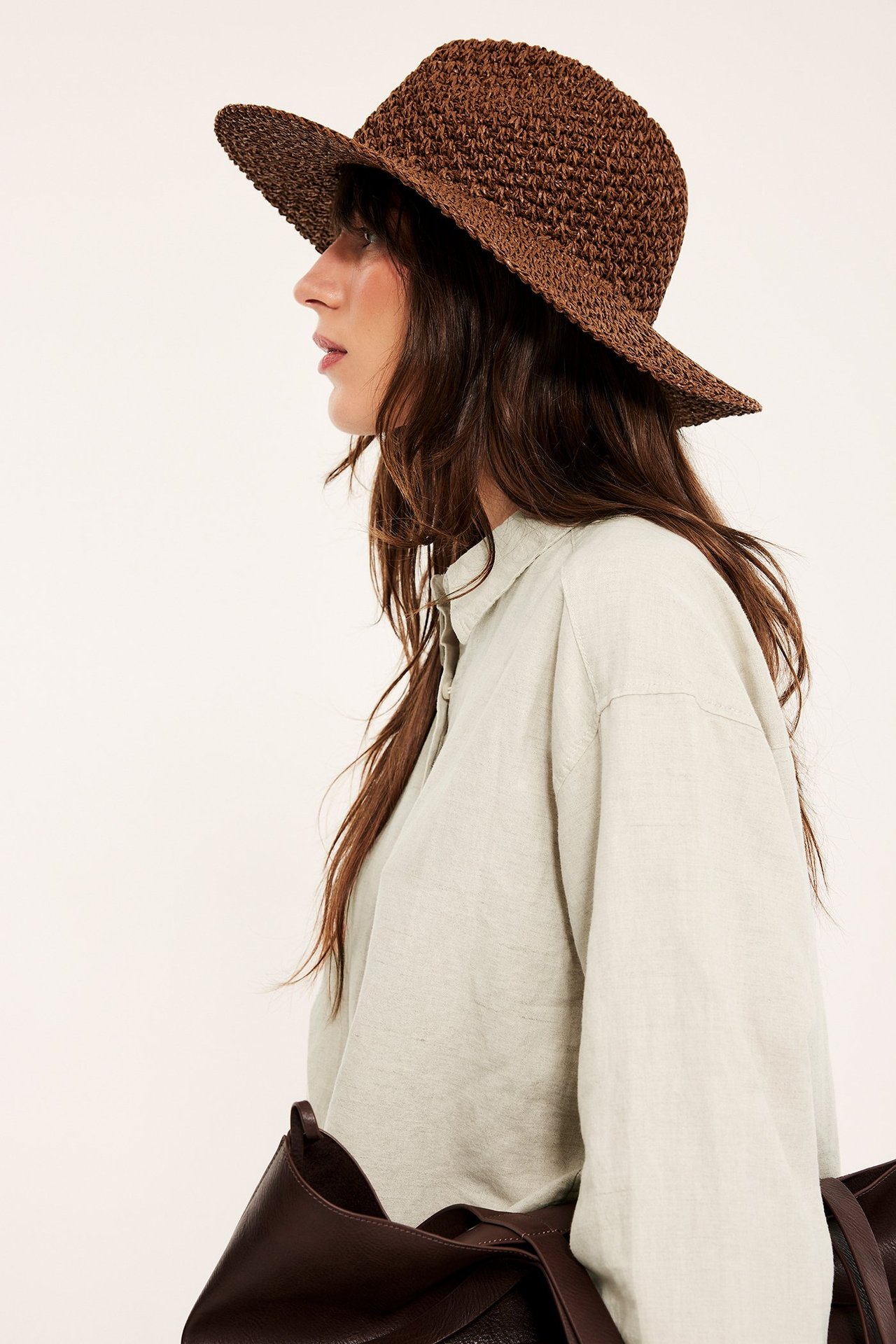 Sivukuva: Nainen yllään ylisuuri beigemelerattu pellavapaita, ruskea hattu, kantaen ruskeaa laukkua.