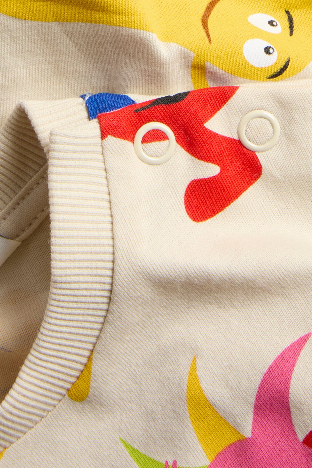 Närbild av en ljusbeige barnt-shirt i bomull med färgglatt Babblarna-tryck och vita tryckknappar.