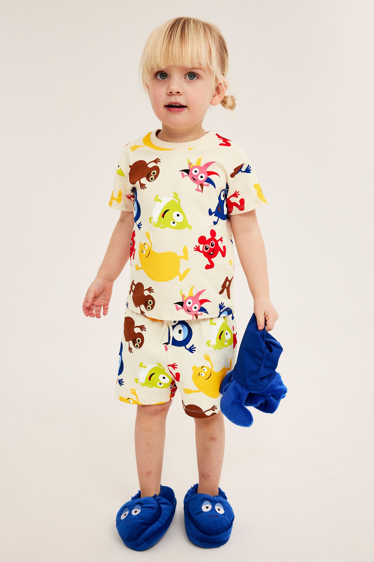 Framifrån: Barn i ljusbeige Babblarna-mönstrad t-shirt och shorts-set, blå tofflor, håller blått mjukisdjur.
