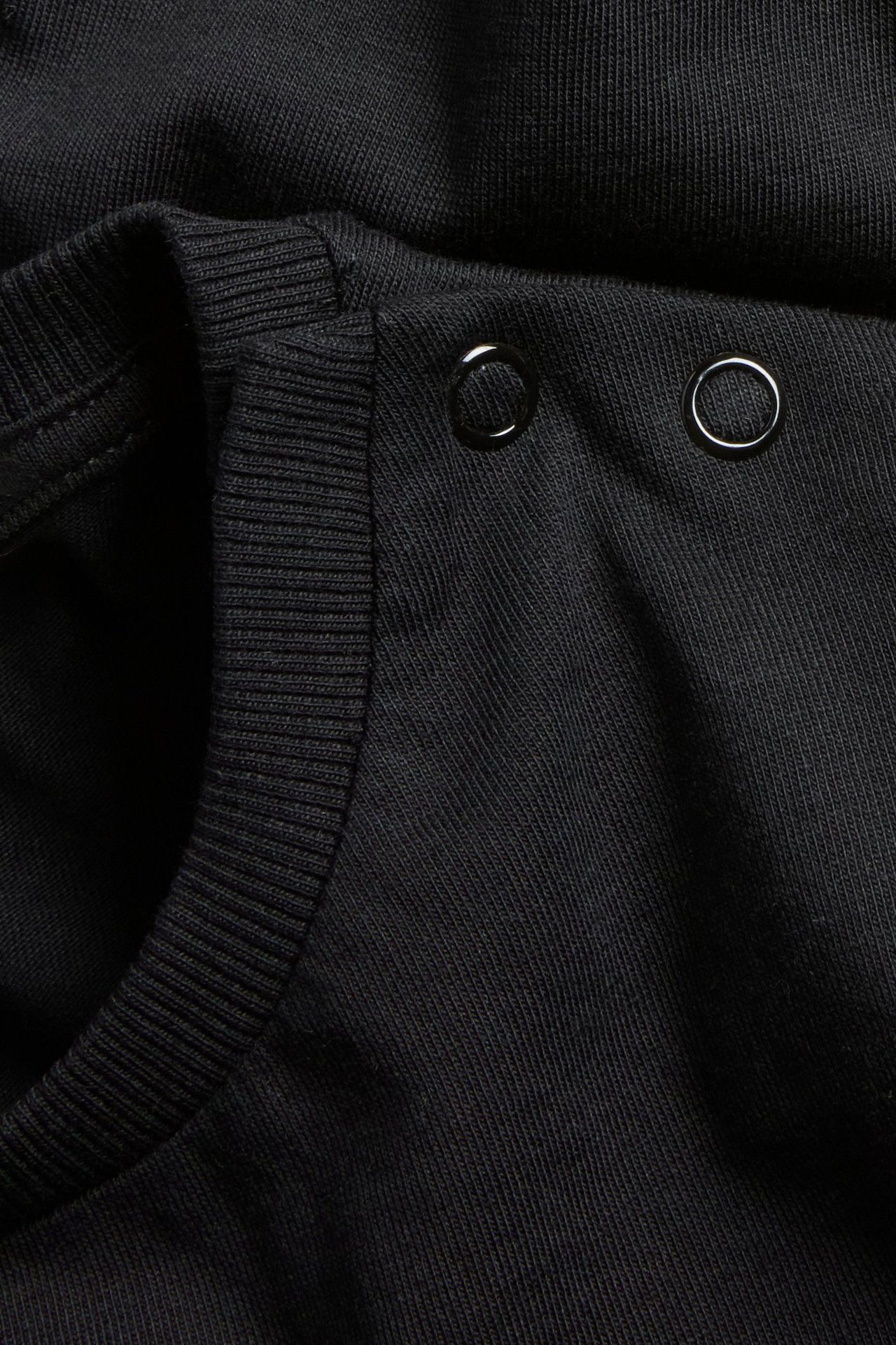 Lähikuva mustasta puuvillaisesta t-paidasta, jossa on rypytetty kaula-aukko ja kaksi metallinepparia.