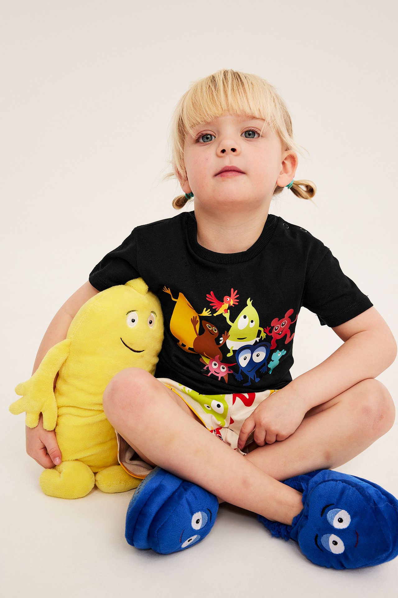 Barn i svart bomull Babblarna t-shirt, mönstrade shorts, blå karaktärstofflor, håller gul mjukisleksak.