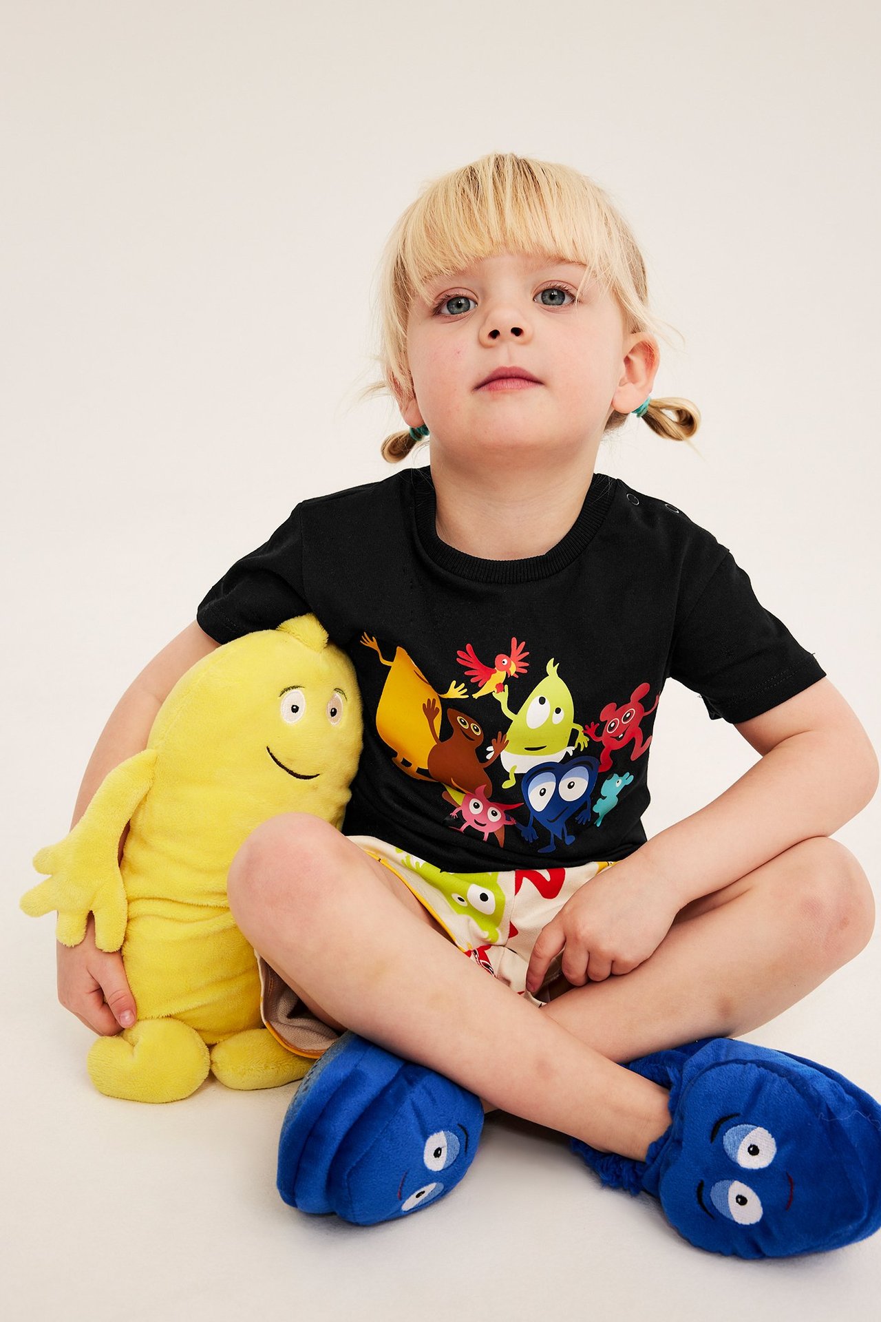 Dziecko w czarnej bawełnianej koszulce Babblarna, wzorzystych szortach i niebieskich kapciach, trzymające żółtą maskotkę.