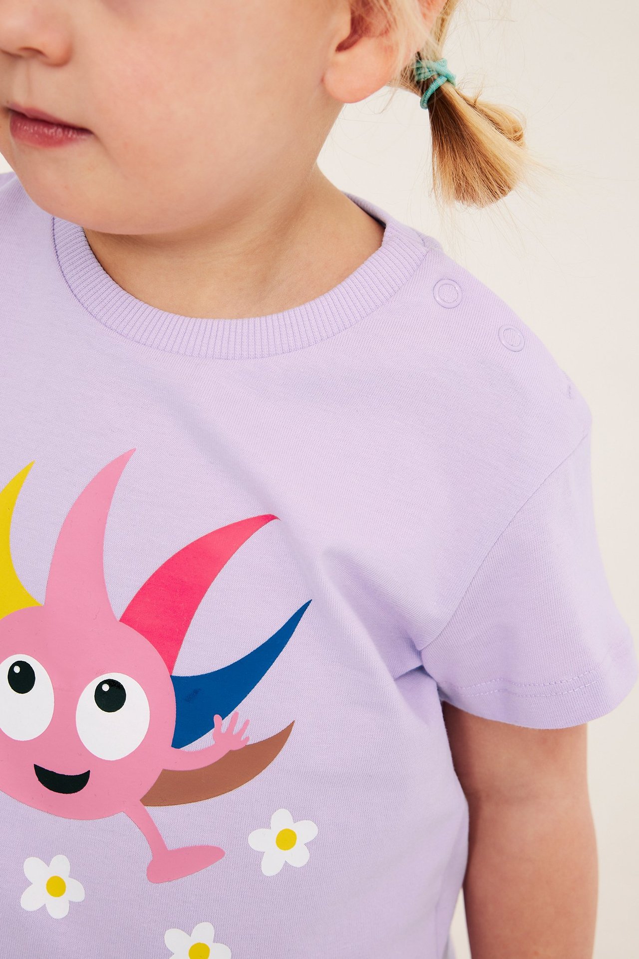 Zbliżenie z przodu: dziecko w liliowym bawełnianym t-shircie z nadrukiem Babblarna i zatrzaskami.