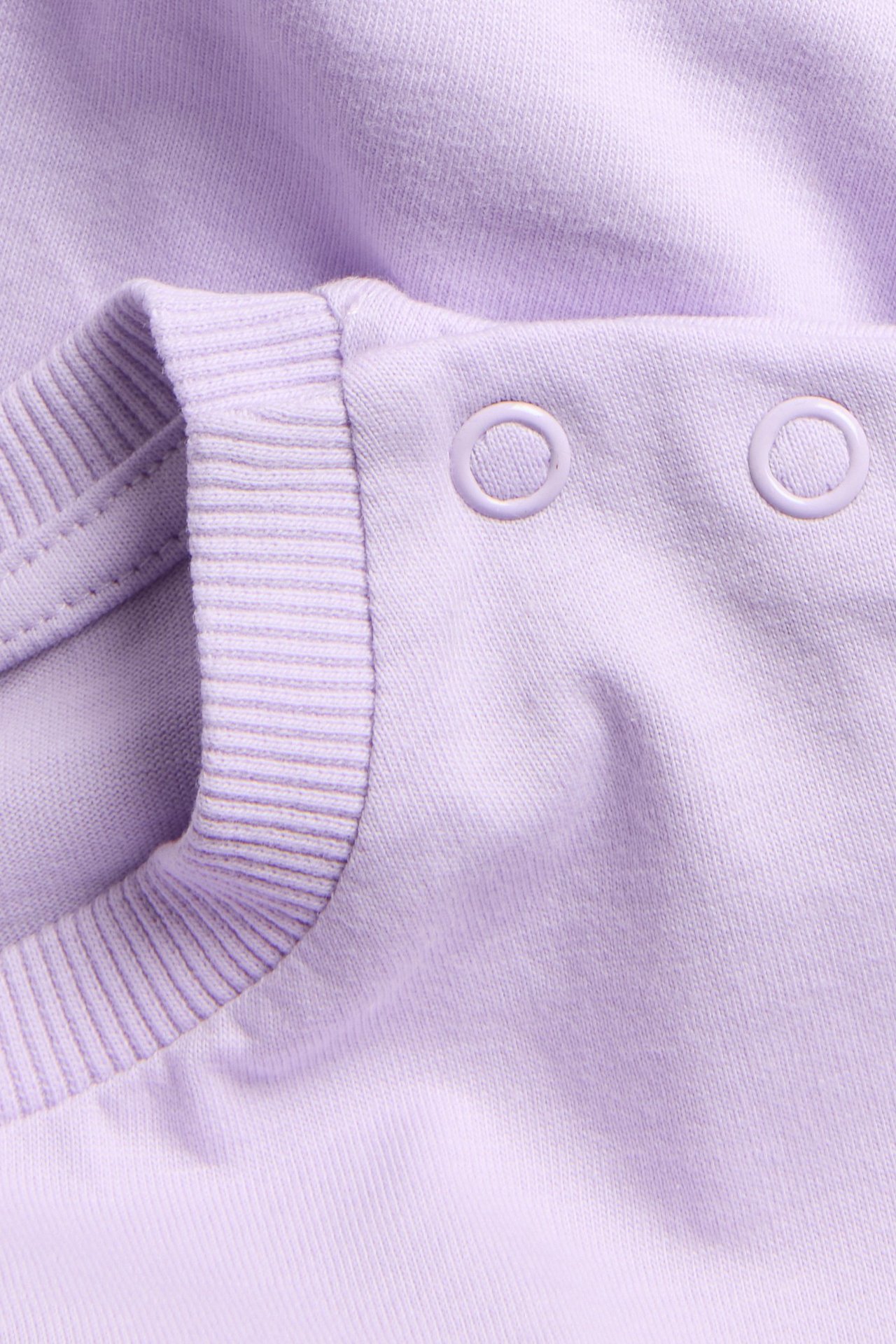 Närbild av en lila bomullst-shirt för barn med ribbad halslinning och två tryckknappar på axeln.