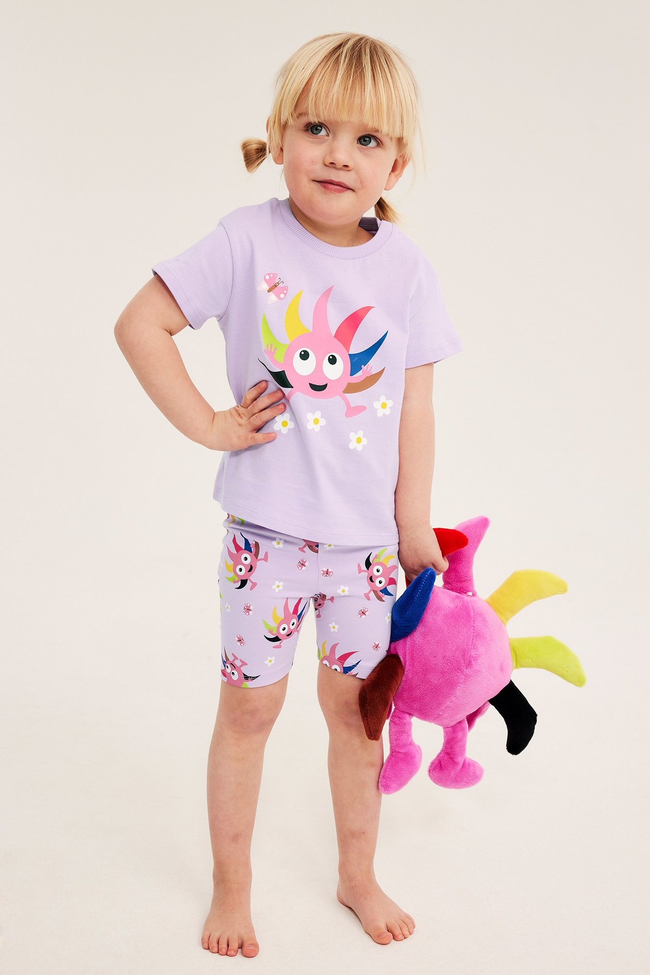 Front view of a kid in a lila Babblarna-tryckt bomulls-t-shirt och matchande shorts, hållandes ett gosedjur.