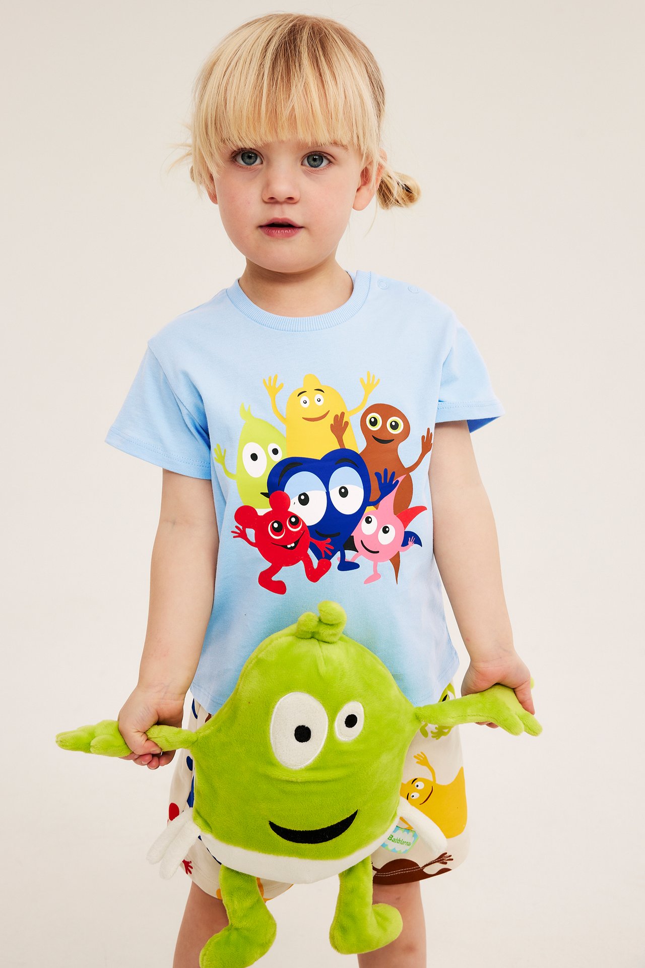 Framifrån: Ett barn i ljusblå bomulls-t-shirt med färgglatt Babblarna-tryck, håller en grön mjukisleksak.