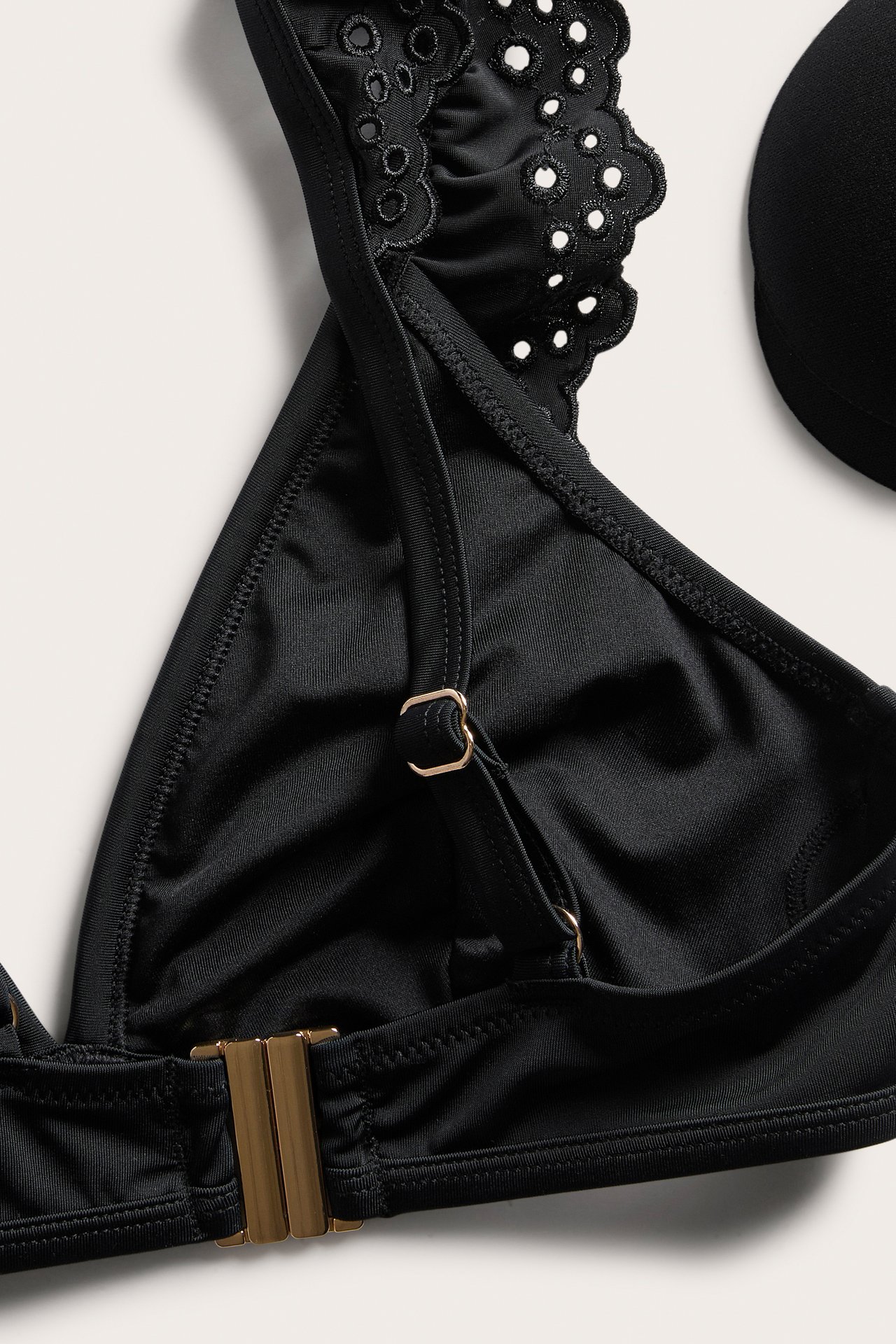 Zbliżenie czarnego topu bikini z falbankami broderie anglaise, złotymi zapięciami i widoczną wkładką.