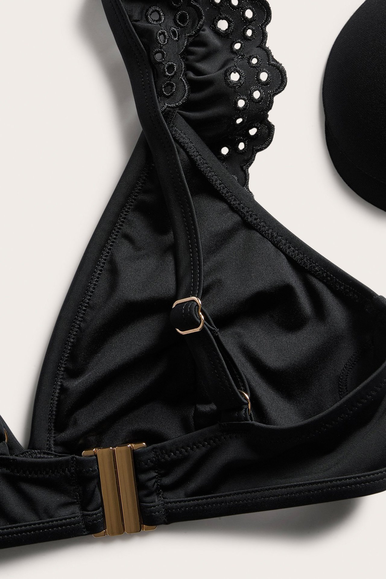 Zbliżenie czarnego topu bikini z falbankami broderie anglaise, złotymi zapięciami i widoczną wkładką.