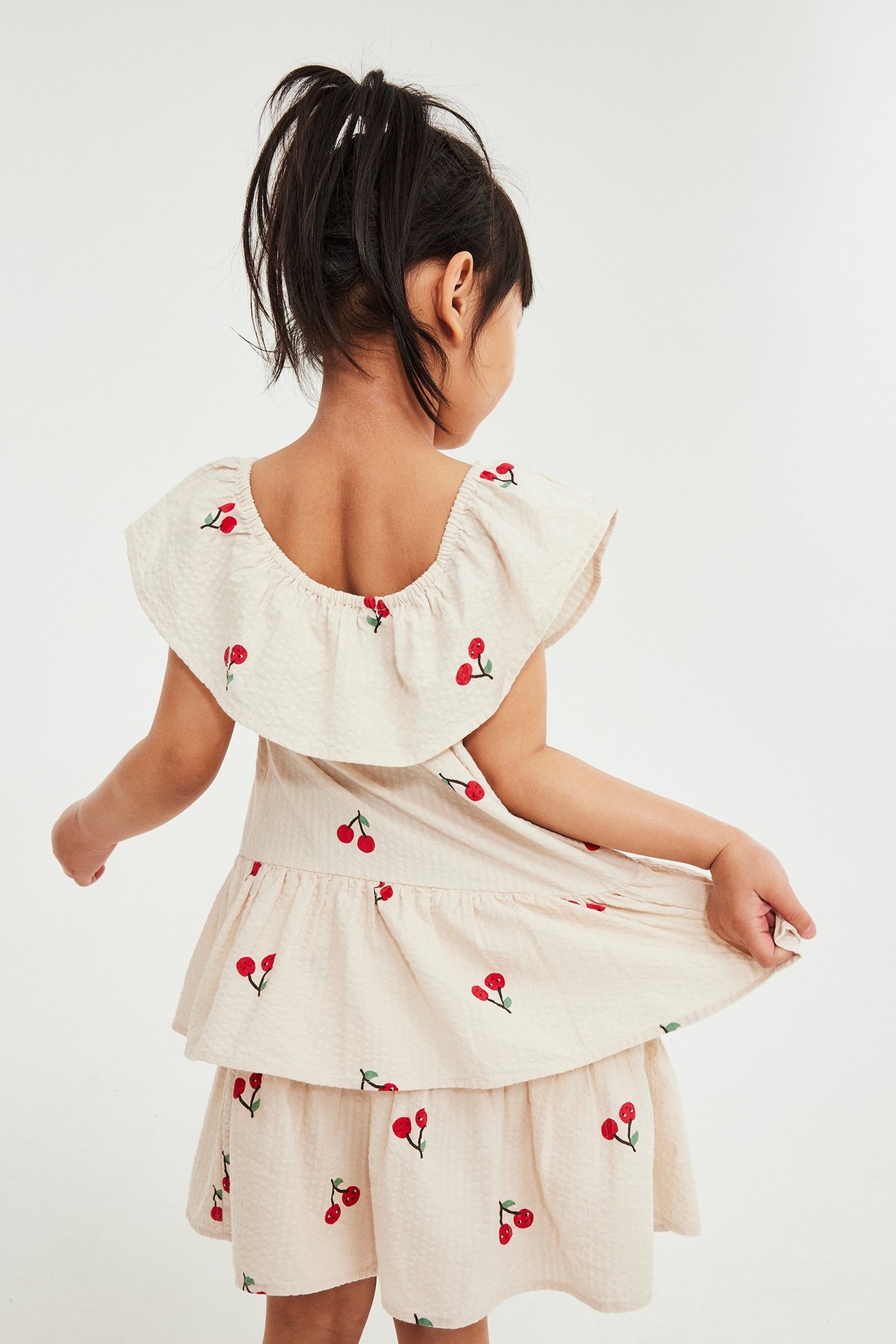 Takakuva: Lapsi beigessä kirsikkakuvioisessa seersucker-mekossa röyhelöillä, pitäen helmaa.