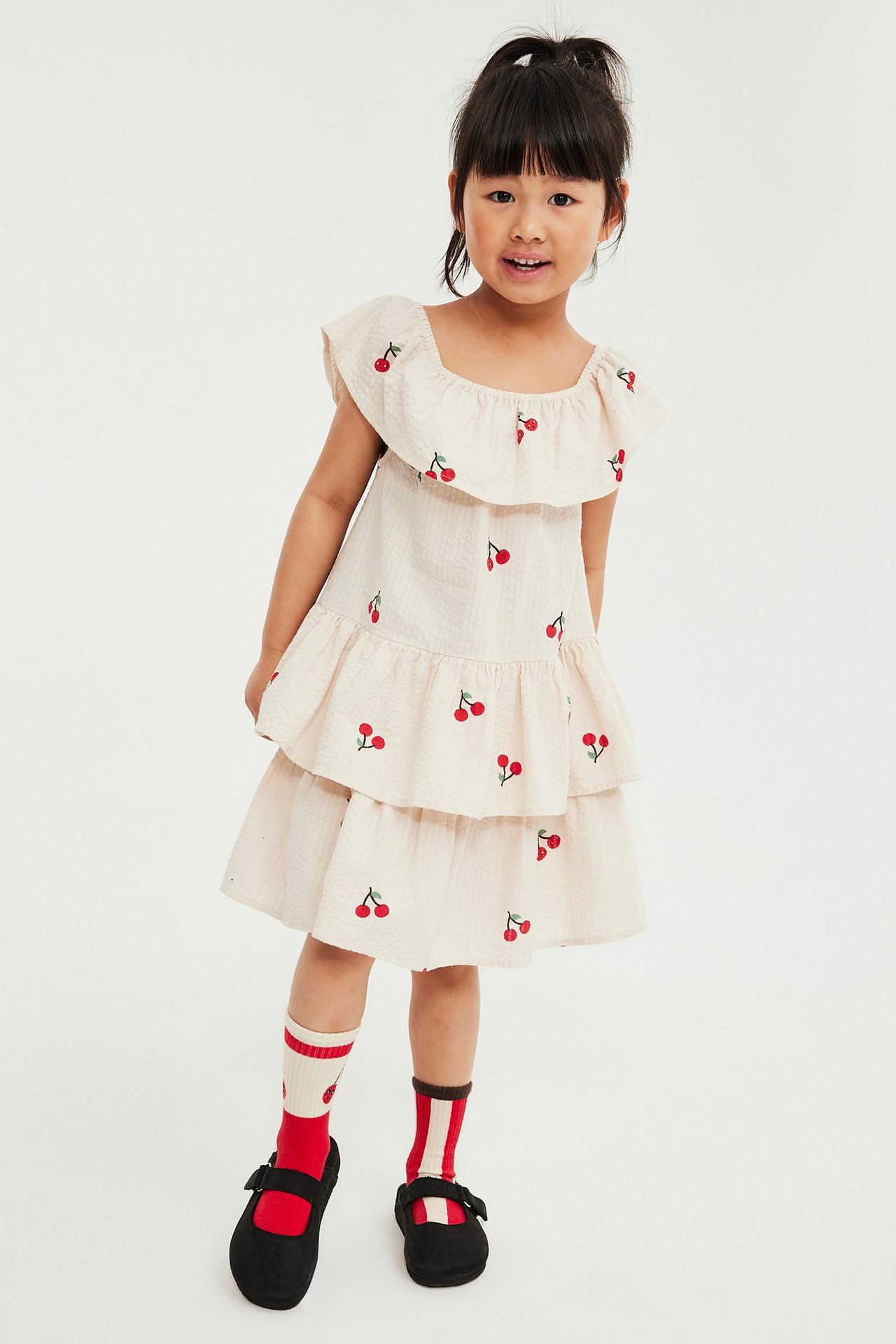 Framifrån: ett barn i beige vävd klänning med körsbärsmönster, mönstrade strumpor och svarta skor.