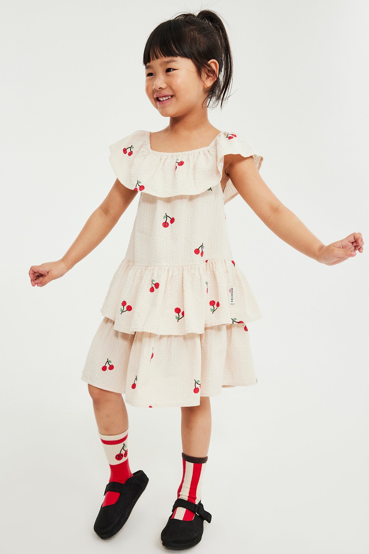 Edestä: Lapsi beigessä seersucker-mekossa kirsikkakuviolla, raitasukissa ja mustissa kengissä.