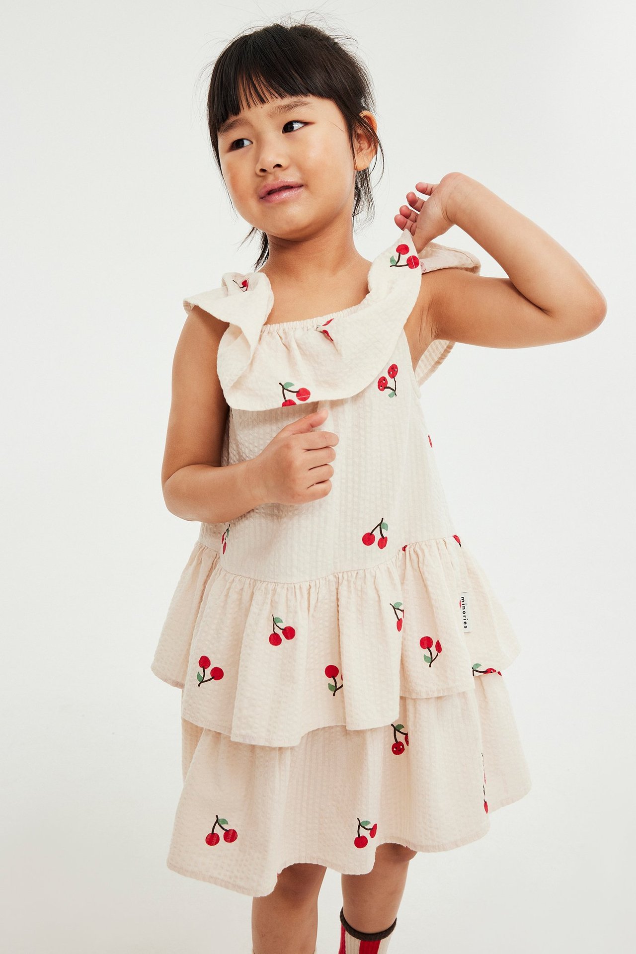 Edestäpäin, lapsi yllään beige seersucker-mekko punaisella kirsikkakuviolla ja röyhelökauluksella.