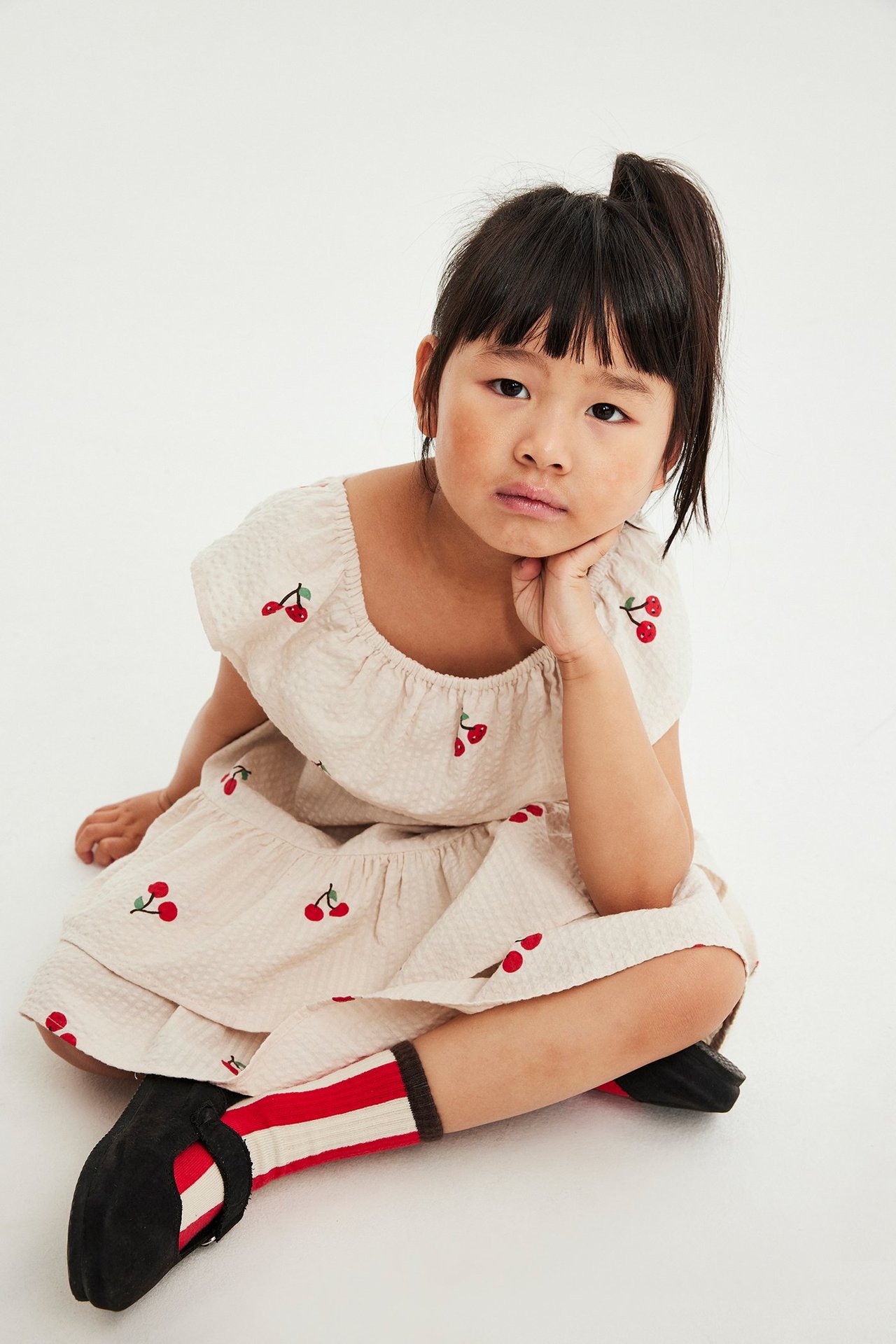 Lapsi, edestäpäin, yllään beige kirsikkakuvioinen seersucker-mekko röyhelöillä ja raidalliset sukat.