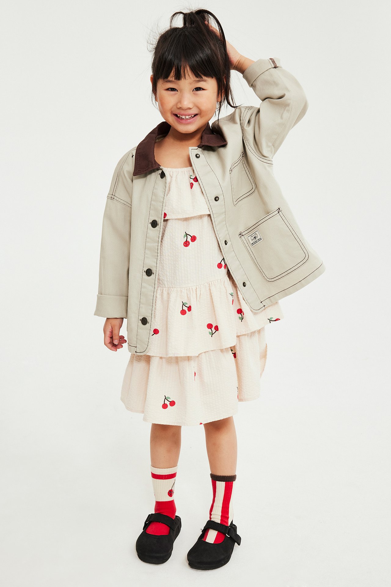 Etukuva lapsesta: beige kirsikkakuvioinen röyhelömekko, vaalea takki, raitasukat ja mustat kengät.