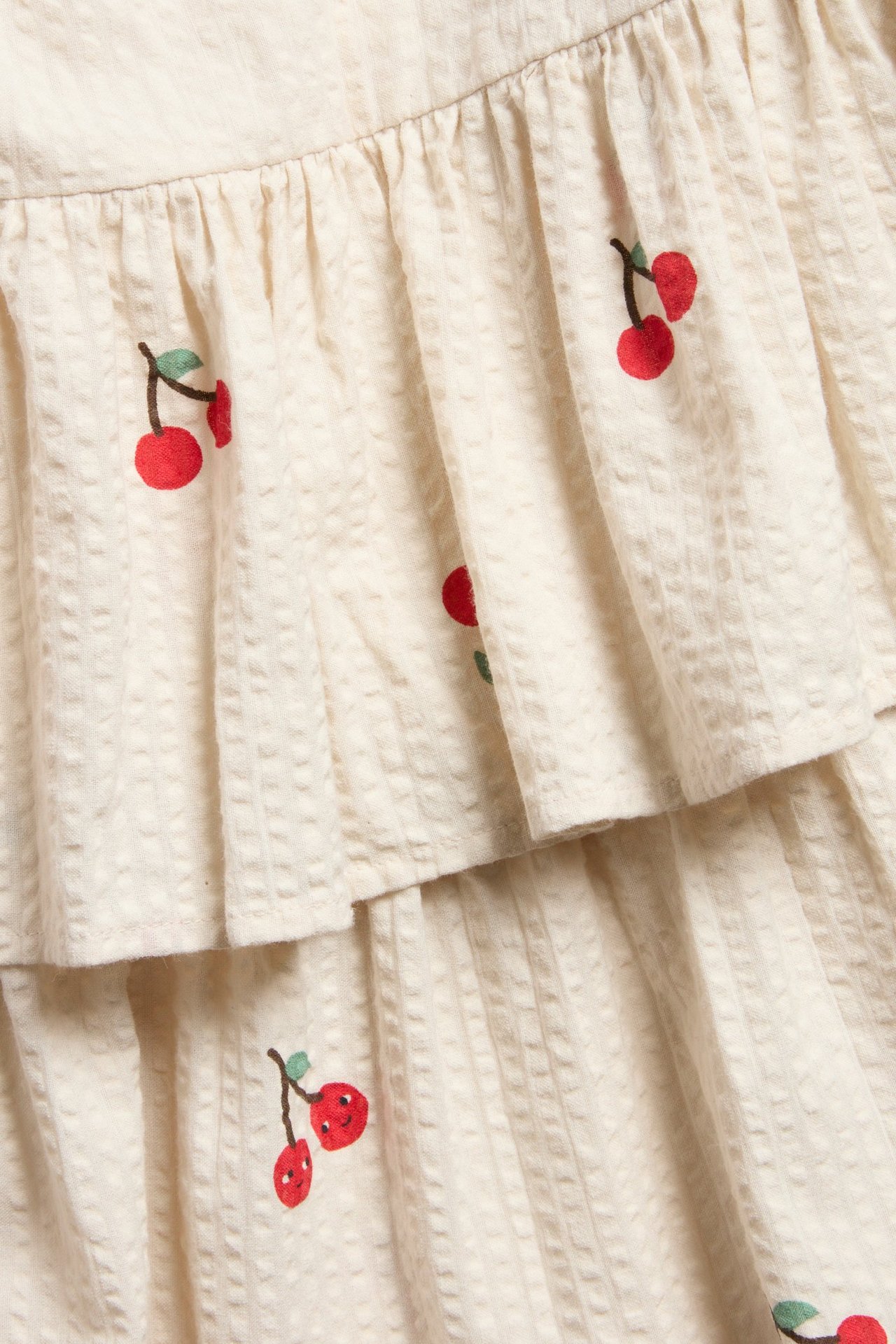 Nærbilde av en beige vevd seersucker-kjole med volanger og rødt kirsebærmønster.