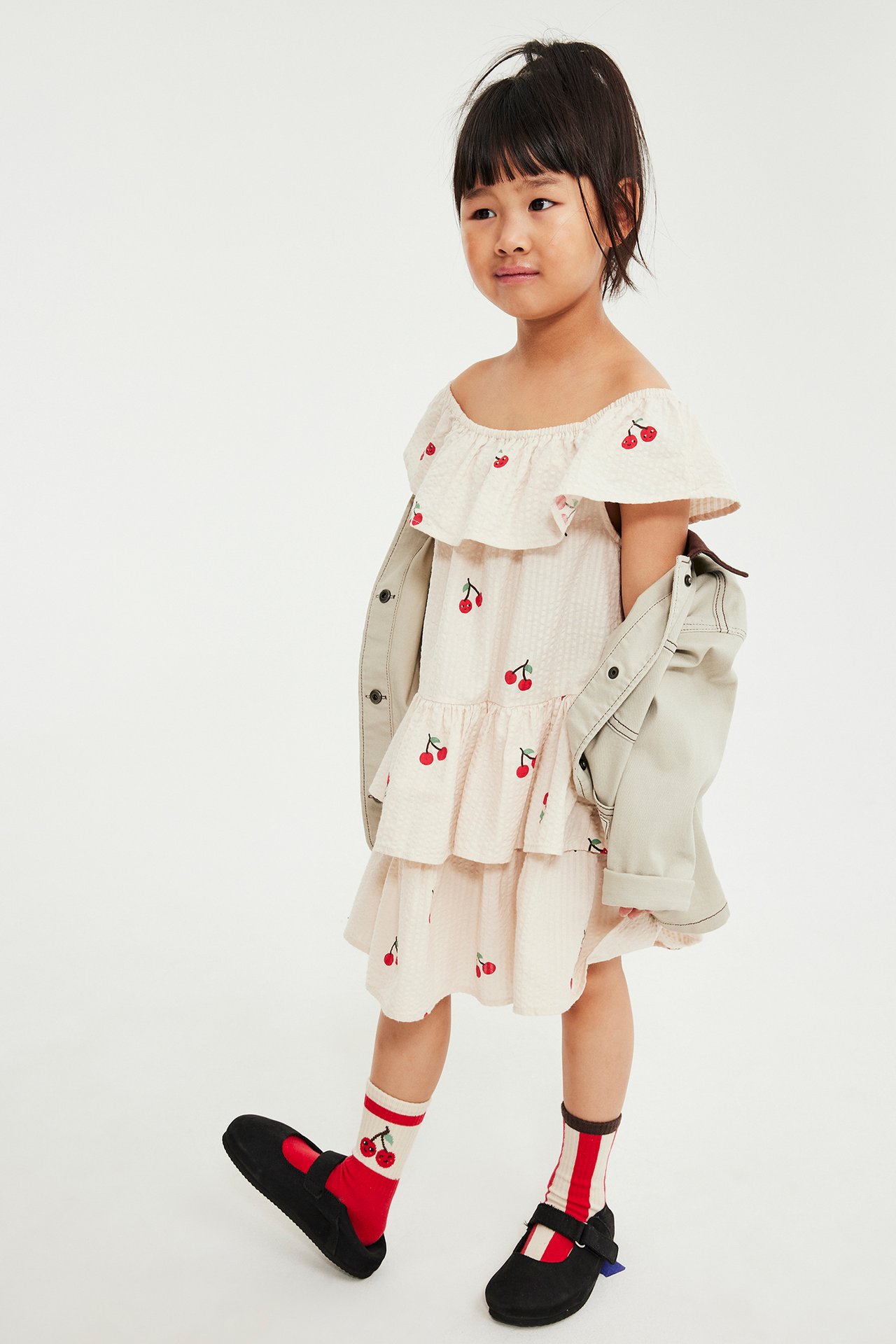 Dziecko w beżowej sukience z falbanami i wiśniami, jasna kurtka na ramionach, czerwone skarpety i czarne buty.