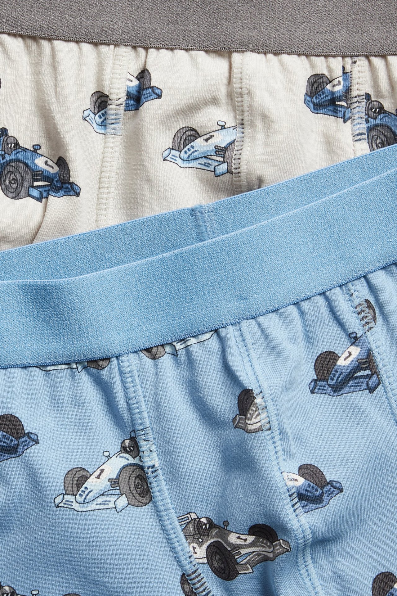 Zbliżenie na dwie pary bokserek dziecięcych: jasnoniebieskie z niebieskim pasem, jasnobeżowe z szarym, obie w wyścigowy wzór.