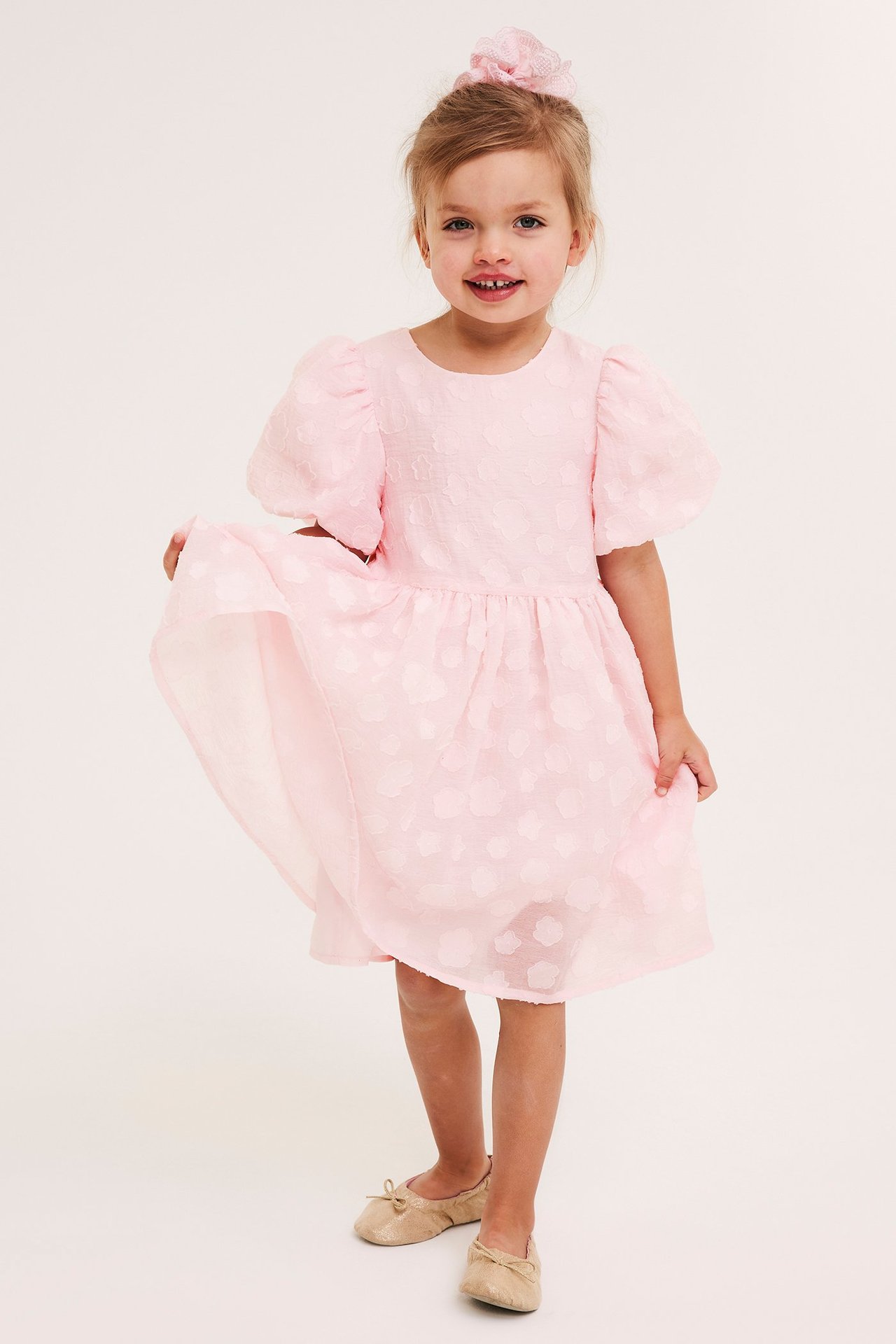Framifrån: Ett barn i ljusrosa organzaklänning med puffärmar, 3D-blommor och beiga ballerinaskor.