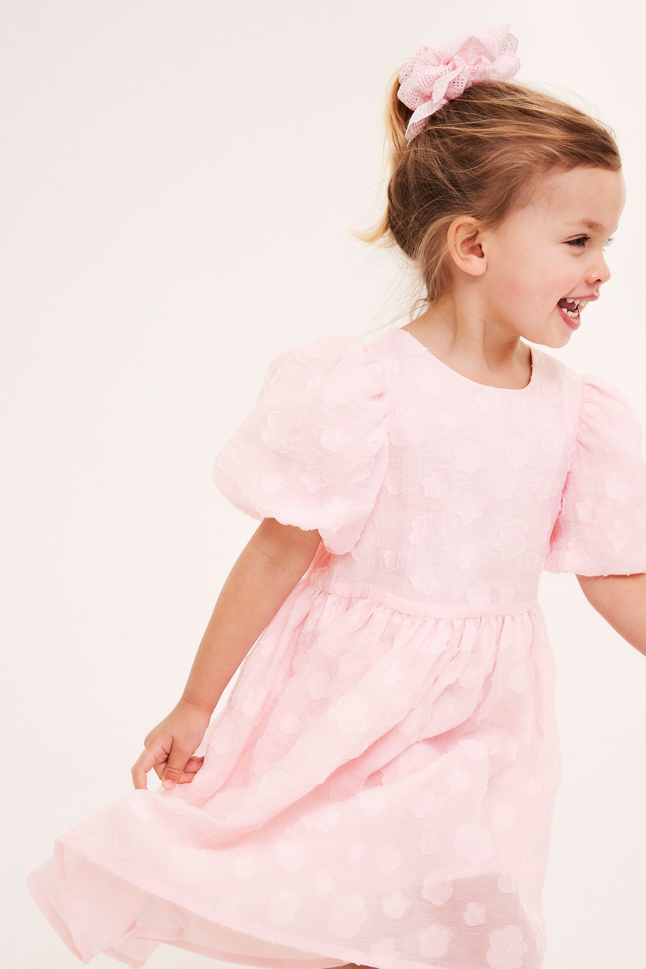 Sidovy: Ett barn i en ljusrosa organzaklänning med puffärmar, texturerade blommor och en matchande scrunchie.