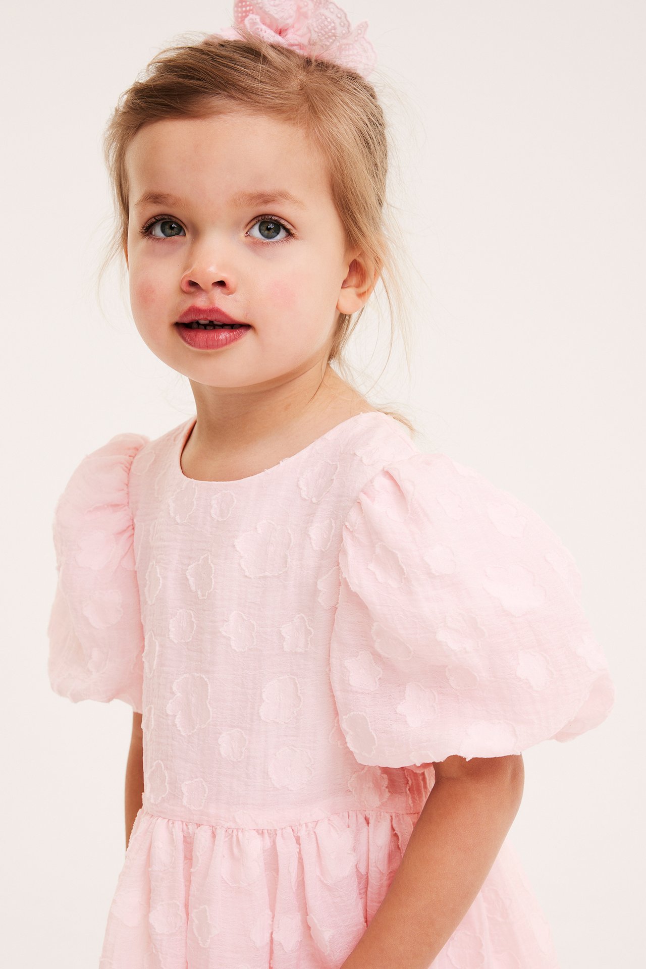 Frontvy av ett barn i en ljusrosa organzaklänning med 3D-blommor, puffärmar och rosa rosett.