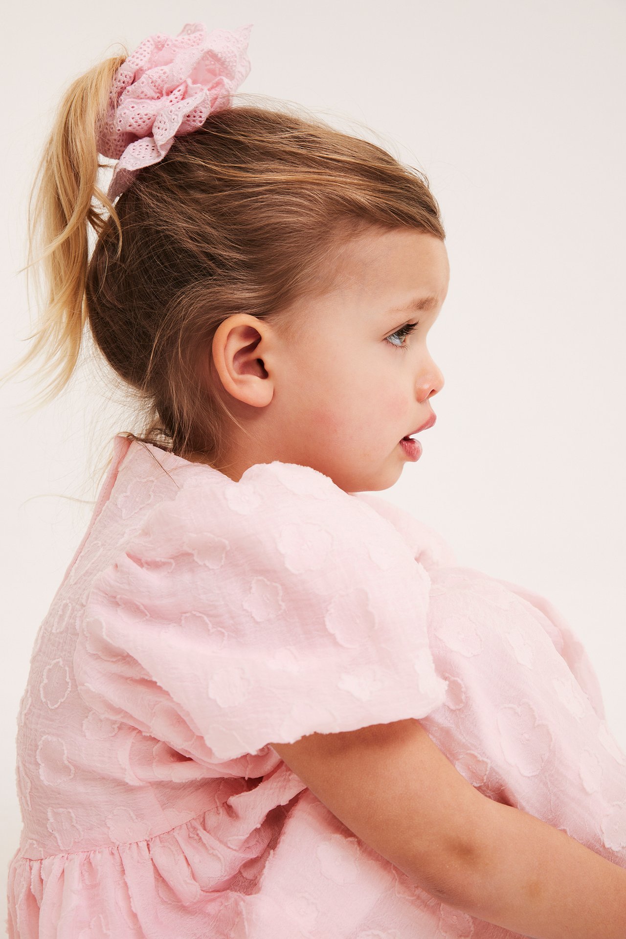 Sidovy av ett barn i en ljusrosa organzaklänning med puffärmar, 3D-blommor och matchande scrunchie.