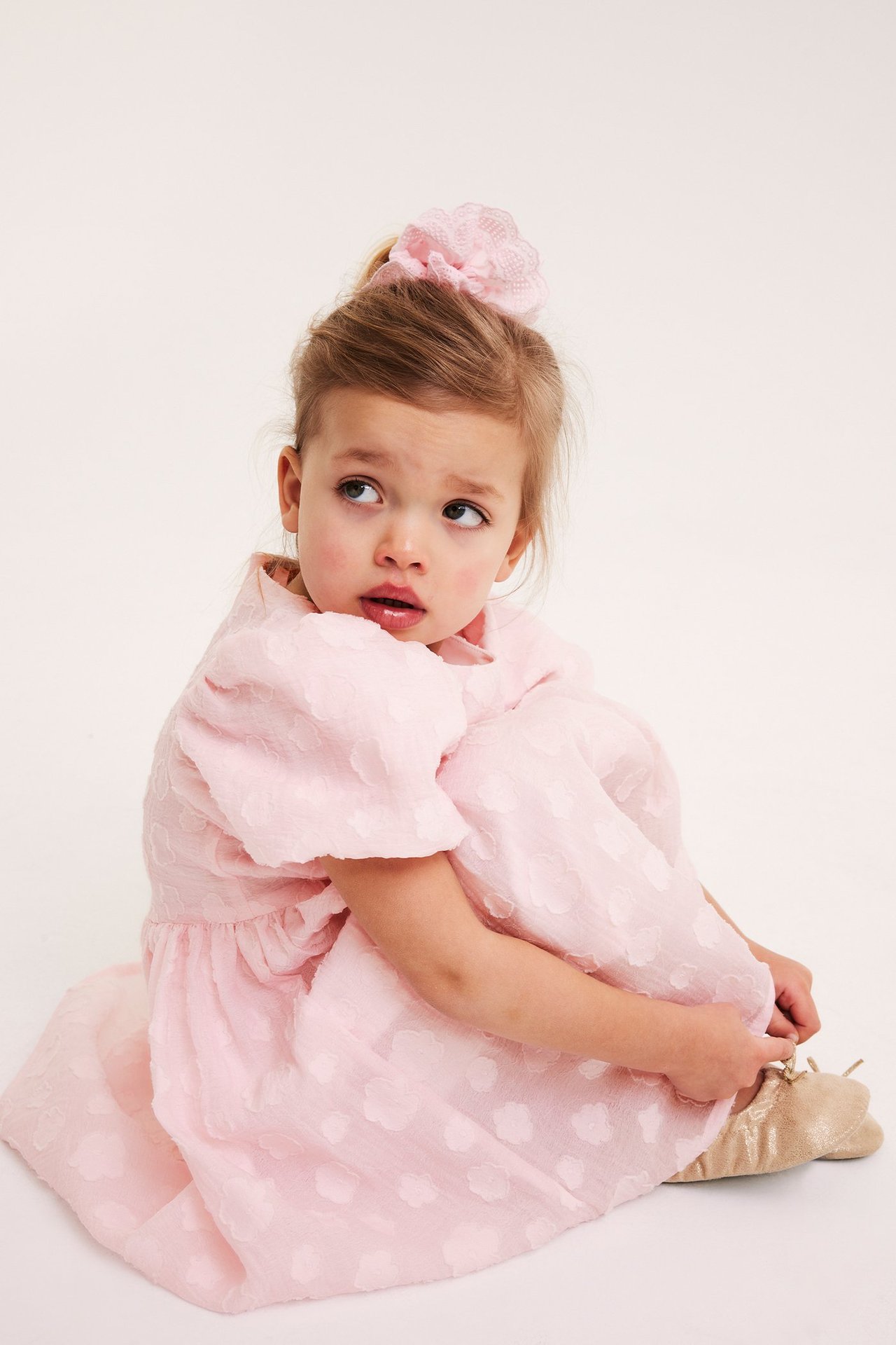 Barn i en ljusrosa organzaklänning med puffärm, blomtextur, rosa scrunchie och guldiga ballerinaskor.
