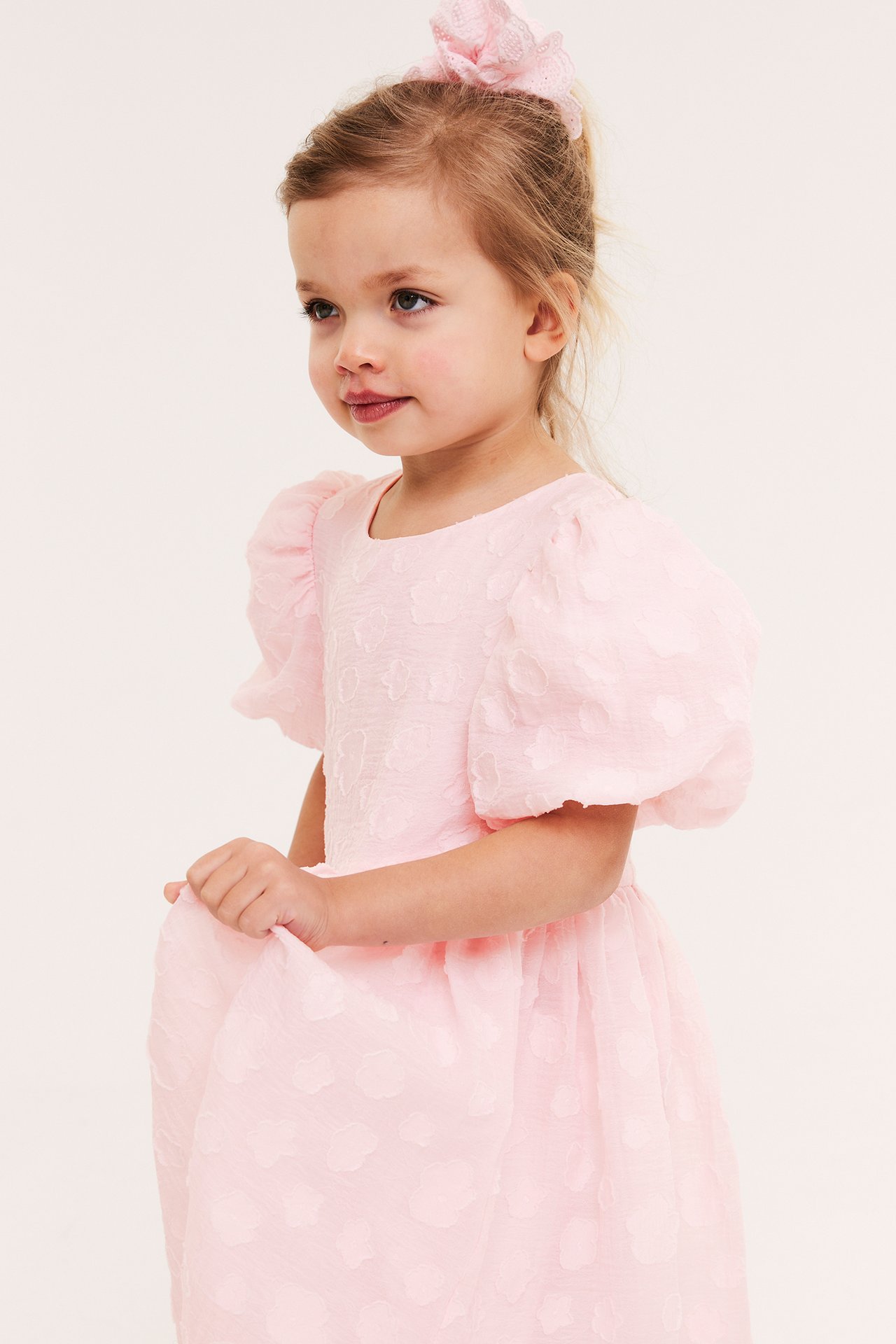 Framifrån: barn i ljusrosa organzaklänning med puffärm, 3D-blommor och matchande hårtofs.