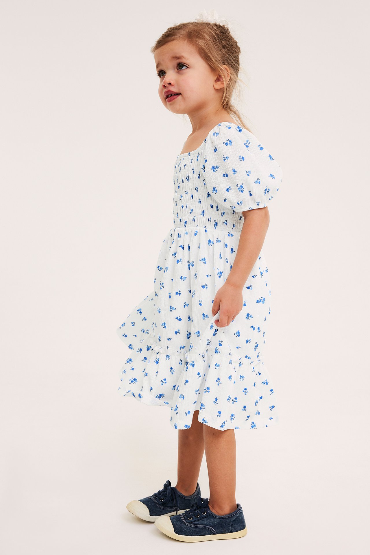 Sidebilde av et barn i en kremfarget blomstret smock-jerseykjole med puffermer og marineblå sneakers.