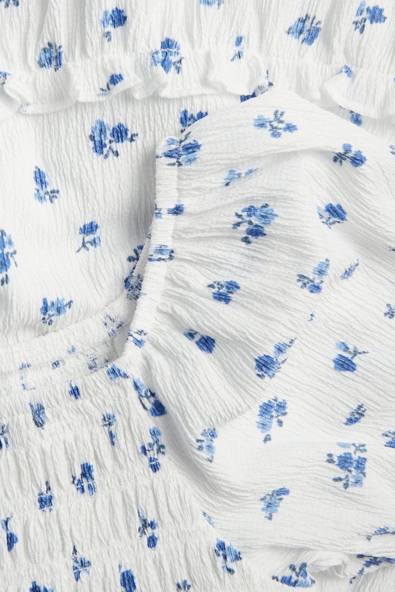 Zbliżenie na kremową sukienkę z marszczonej dzianiny w niebieskie kwiaty, ze smockowymi detalami.