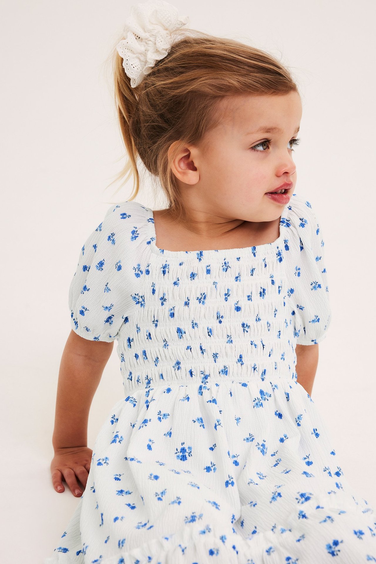 Barn har på seg off-white blomstret krøllet jerseykjole med smocket liv og hvit blondeskrunchie.