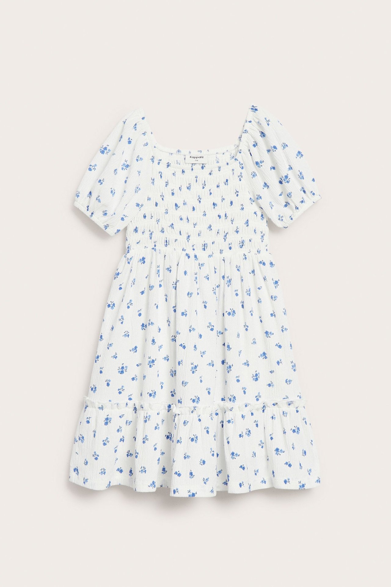 Front: Hvit barnekjole i blomstret jersey med smocket topp, korte puffermer og lagdelt skjørt.