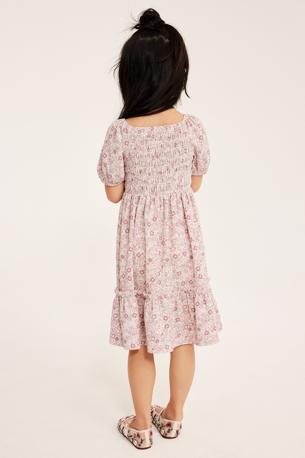Bakifrån på ett barn i en ljusrosa blommig jerseymönstrad klänning med smockat liv och broderade ballerinaskor.