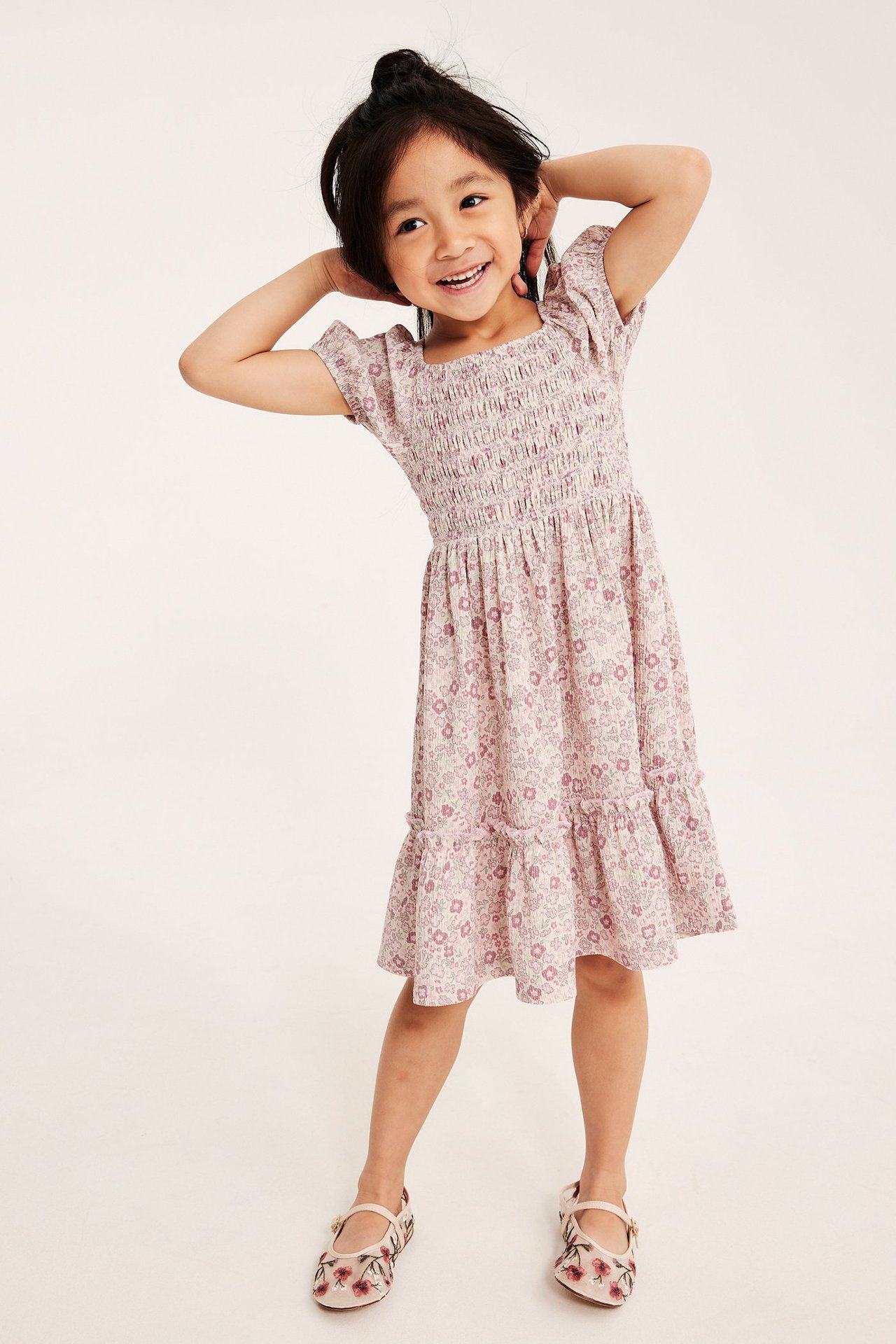 Forfra: barn i lys rosa blomstret jerseykjole med smock-liv og matchende broderte sko.