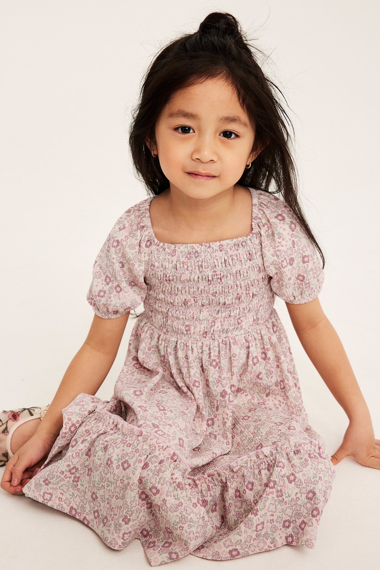 Widok z przodu: siedzące dziecko w jasnoróżowej, kwiecistej sukience z krepowanej dzianiny, ze smockiem i bufiastymi rękawami.