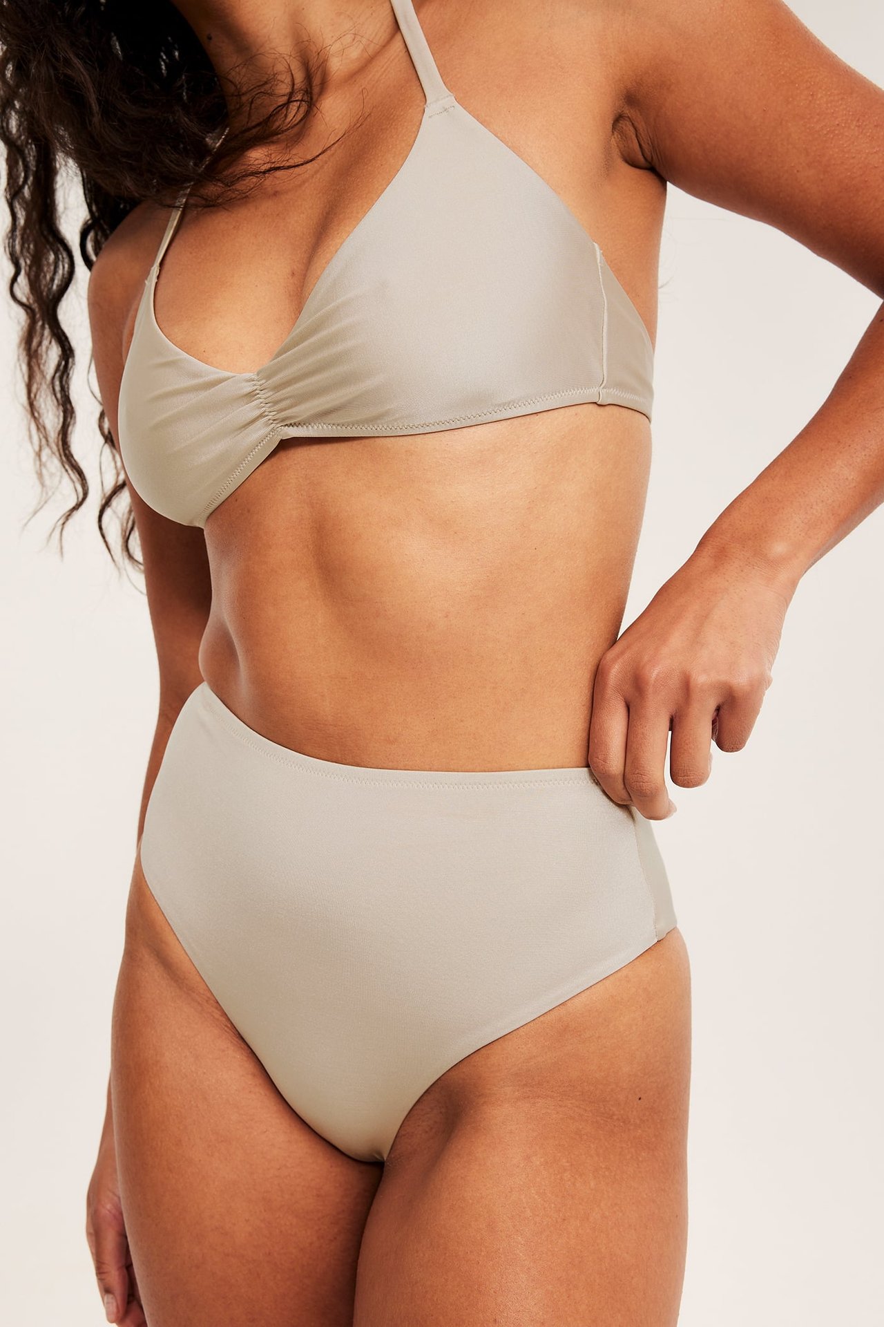 Edestä: nainen yllään beige korkeavyötäröinen brazilian-bikinihousu, yhteensopiva rypytetty halterneck-toppi.